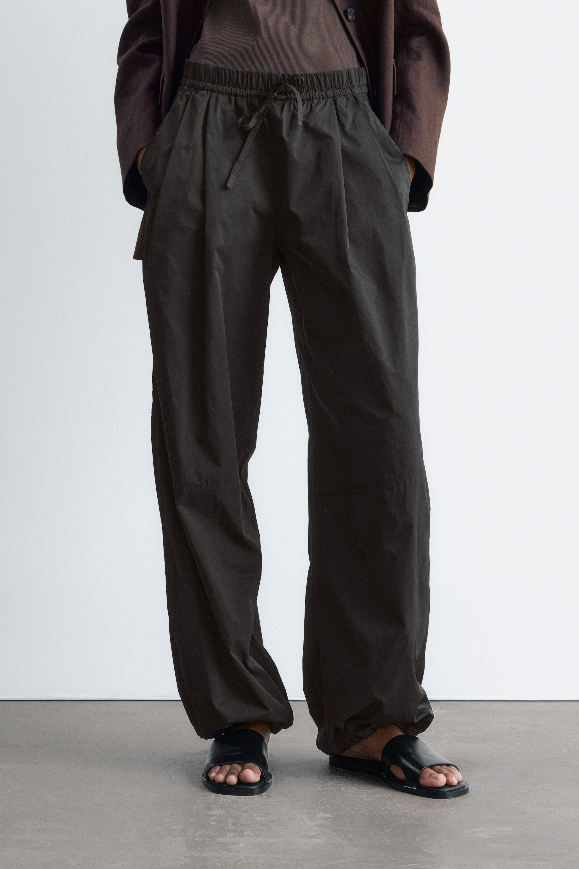 Grösseres Bild ansehen: Dunkle Barrel-Leg-Hosen werden vom Model getragen, mit Frontfalten, einem elastischen Kordelzugbund, der zugebunden ist, und Schnittlinien-Nähten am Knie. Die Hände des Models ruhen in den Seitentaschen, und die Hosen haben elastische, geraffte Säume.