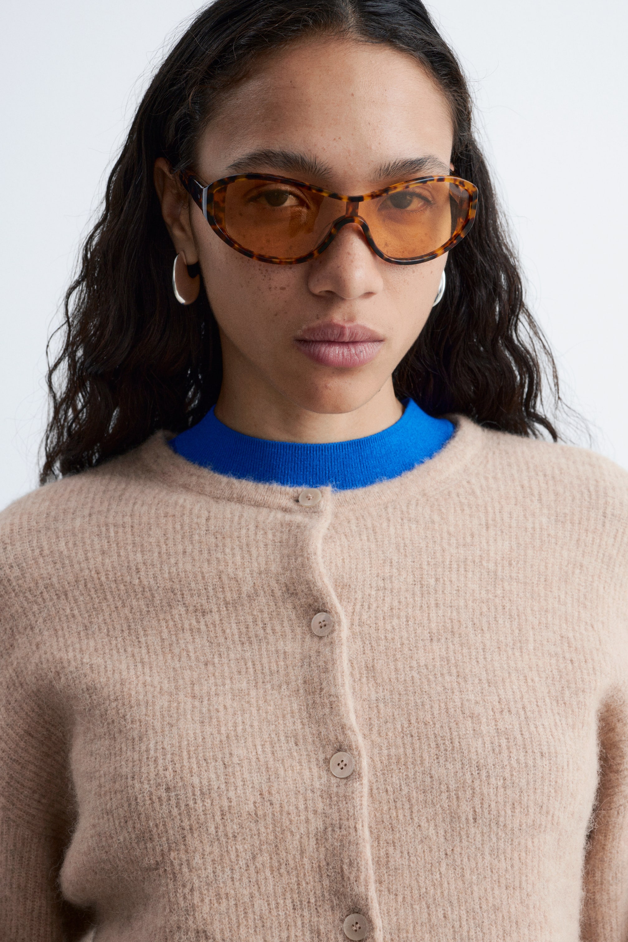 Größeres Bild ansehen: Ein beige-flauschiger Strick-Cardigan mit Rundhalsausschnitt und Knopfleiste mit passenden Knöpfen. Er wird offen über einem blauen Oberteil präsentiert.