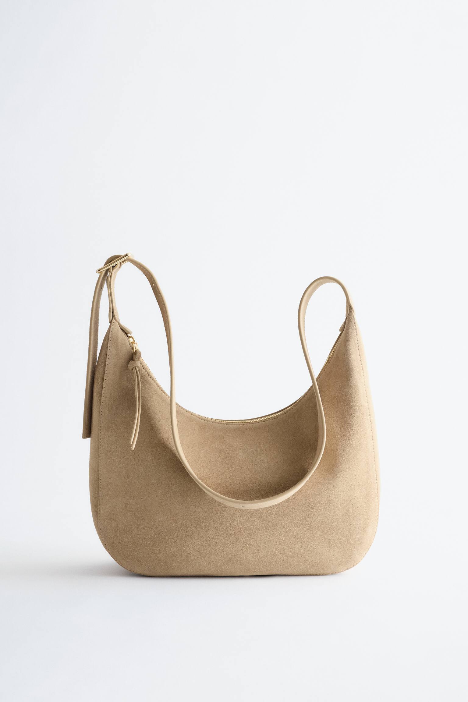 Eine hellbraune Hobo-Tasche mit Wildlederstruktur präsentiert sich von vorne und zeichnet sich durch eine geschwungene, lässige Silhouette aus. Sie verfügt über einen langen, verstellbaren Schulterriemen mit einer goldfarbenen Schnalle und einem goldfarbenen Reißverschluss oben, der mit einem kleinen Zuglaschen-Akzent versehen ist. - 1