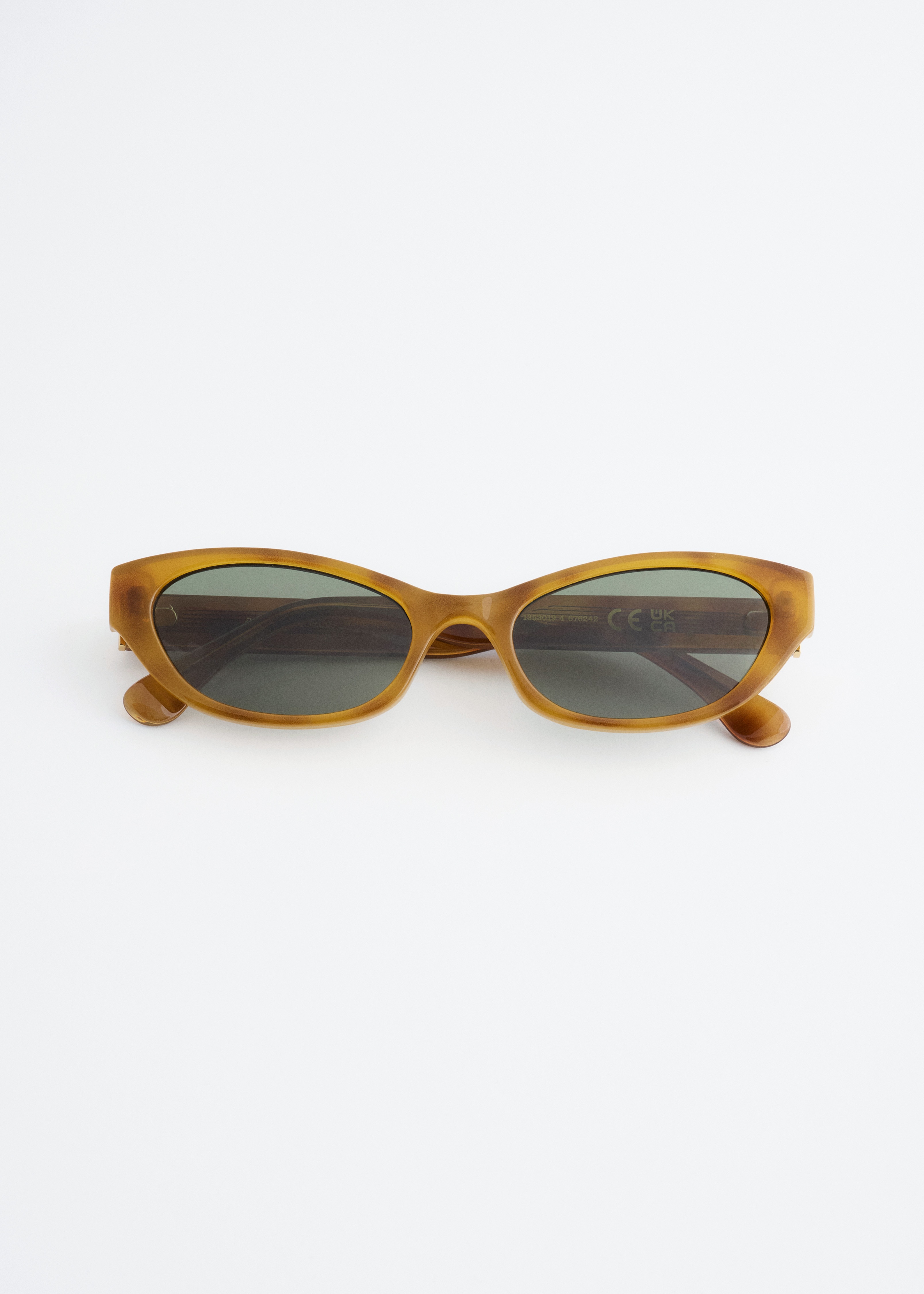 Ãculos de sol estreitos e ovais, com armaÃ§Ãµes translÃºcidas de tom mel e lentes escuras, estÃ£o a ser usados pela modelo. Os Ã³culos de sol combinam com um pequeno brinco de argola dourado e um casaco castanho-escuro.