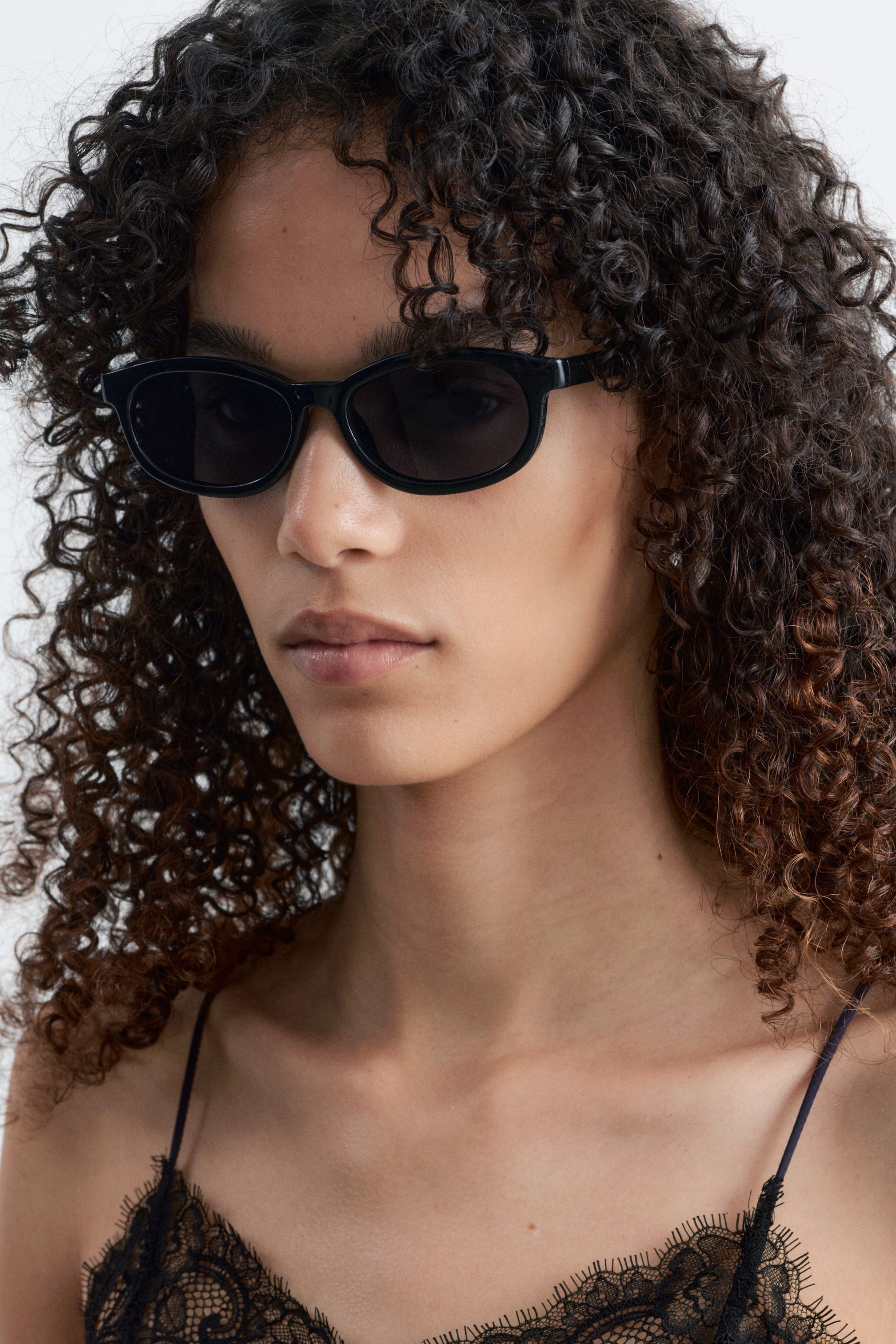 Ver imagem maior: Óculos de sol pretos com armações espessas e suavemente retangulares, equipados com lentes escuras e opacas. Estão a ser usados no rosto de uma pessoa, ligeiramente inclinados para a direita, destacando o seu ajuste e design.