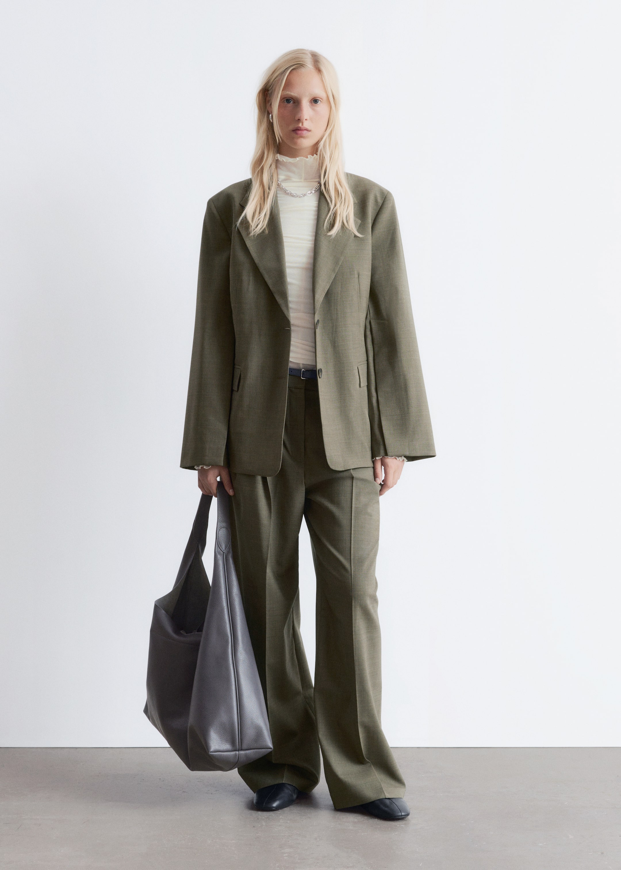 Ingrandisci l'immagine: Un completo verde oliva è messo in mostra da un'angolazione frontale, caratterizzato da un blazer oversize, monopetto con spalle larghe e revers a tacca. Pantaloni a gamba larga abbinati vengono indossati con una cintura scura, e un top leggero, semi-trasparente, a collo alto con bordi arricciati è stratificato sotto il blazer.
