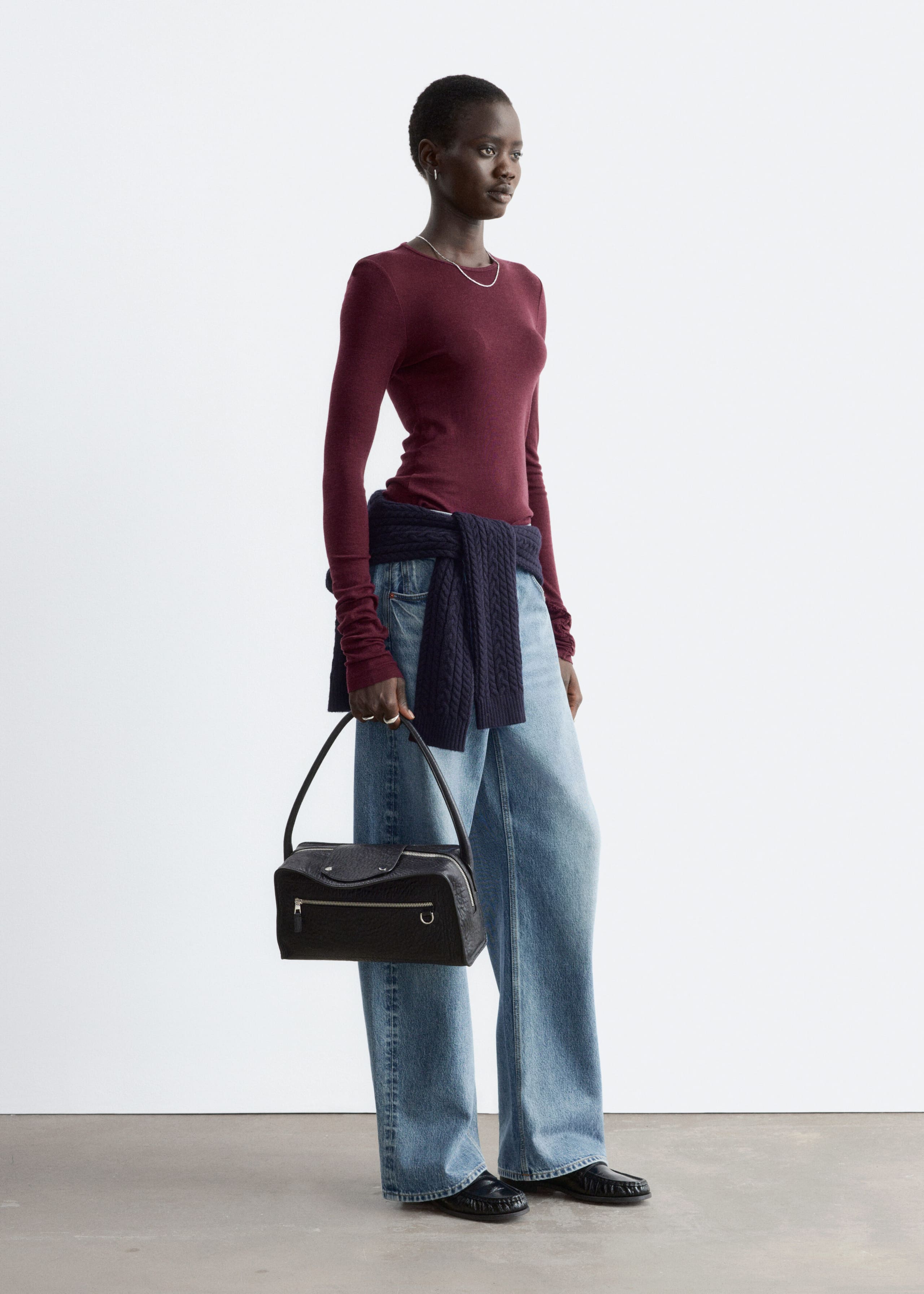 Ein bordeauxrotes, glatt gestricktes Wolloberteil mit Rundhalsausschnitt und langen Ärmeln wird vom Model getragen. Das Oberteil wird mit weit geschnittenen blauen Jeans, einem dunkelblauen Pullover, der um die Taille gebunden ist, und einer schwarzen Lederhandtasche kombiniert, die das Model in der Hand hält.