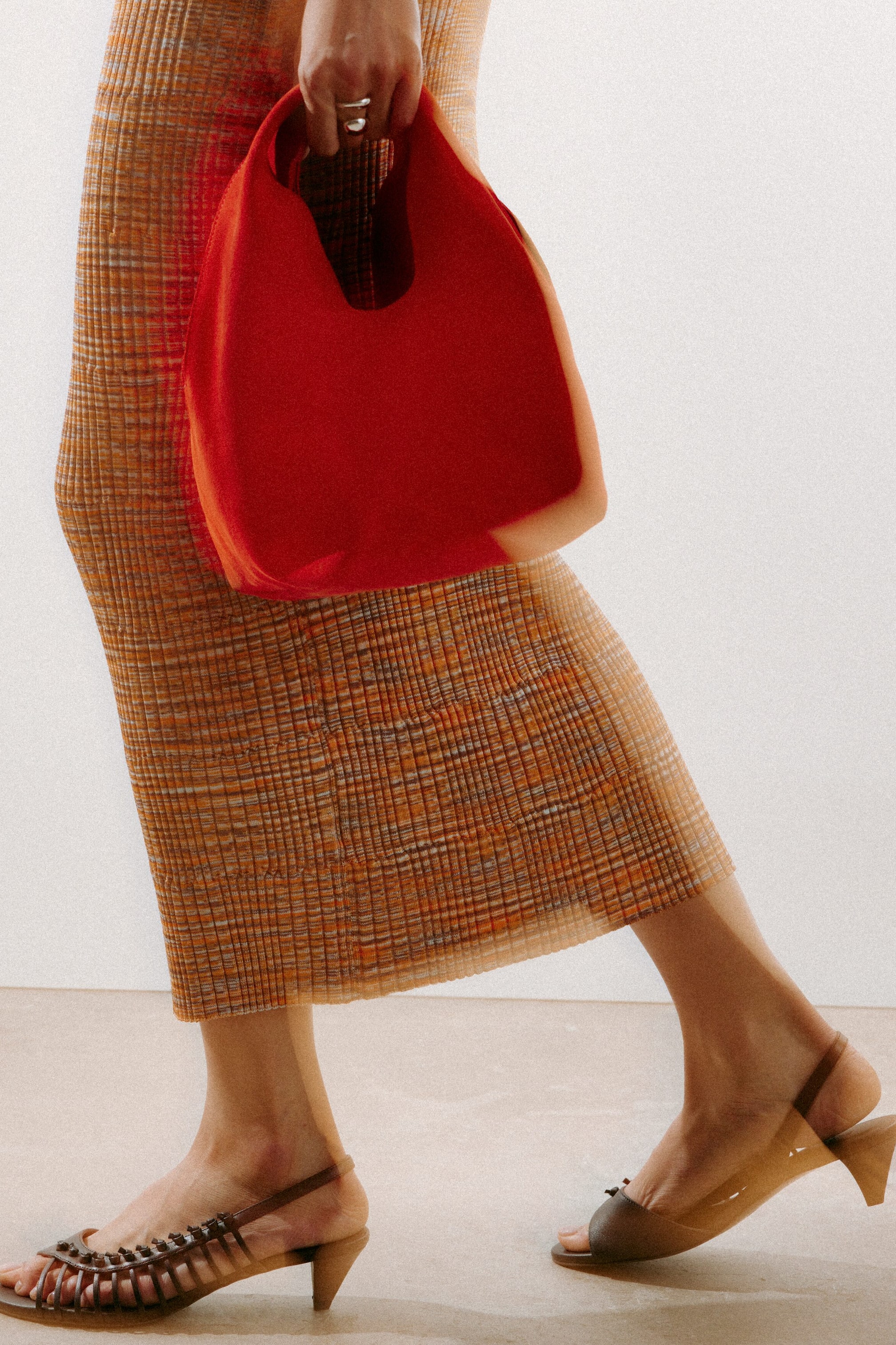 Zobrazit větší obrázek: Matná červená, nestrukturovaná taška je držena v ruce, čímž vyniká její měkký, zakřivený tvar a integrované výřezy pro držadlo.