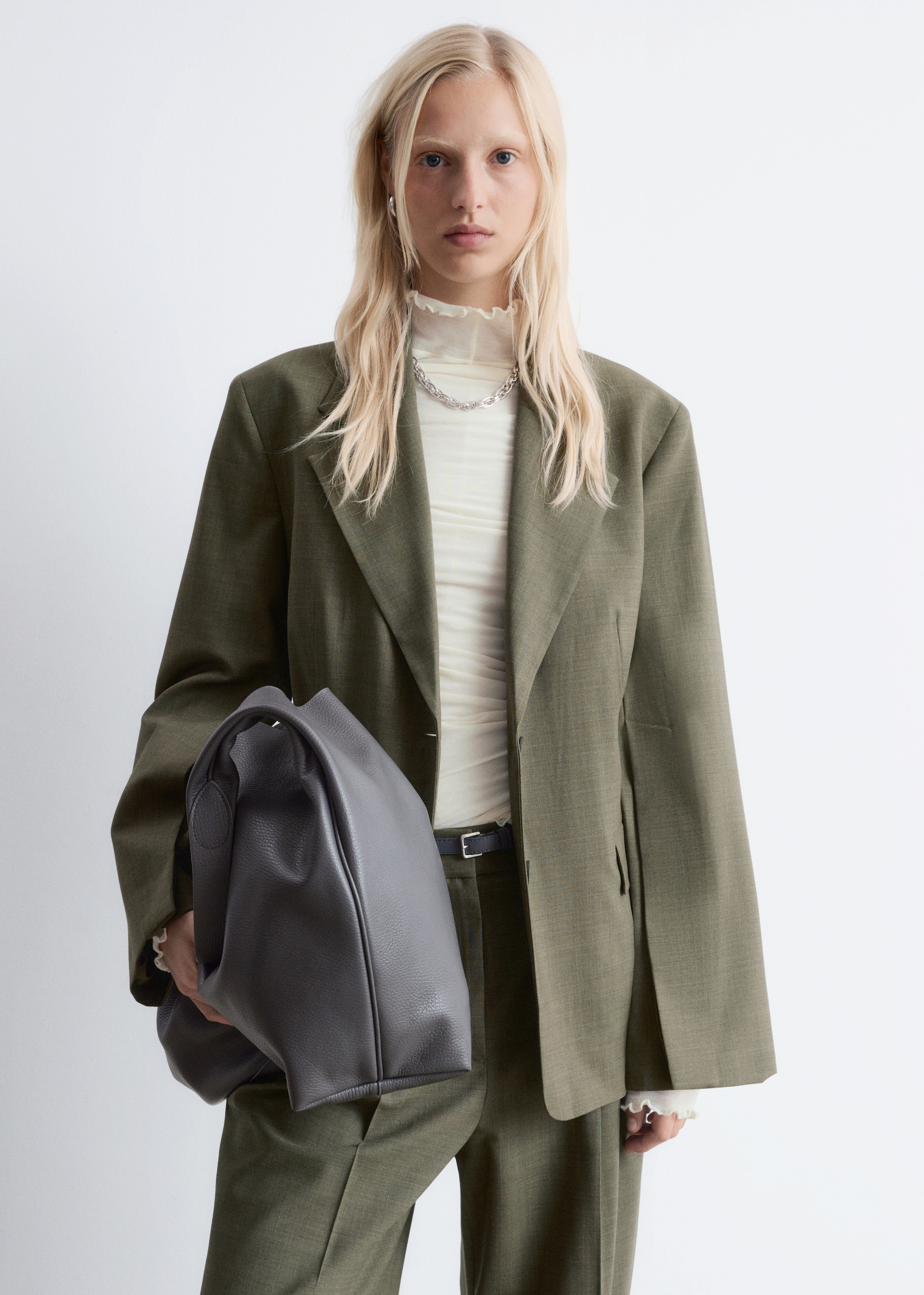 Ingrandisci l'immagine: Un blazer verde oliva, realizzato in un tessuto intrecciato e strutturato, è indossato. Presenta rever a dente e una vestibilità comoda, con una maglia a balze di colore chiaro sotto.