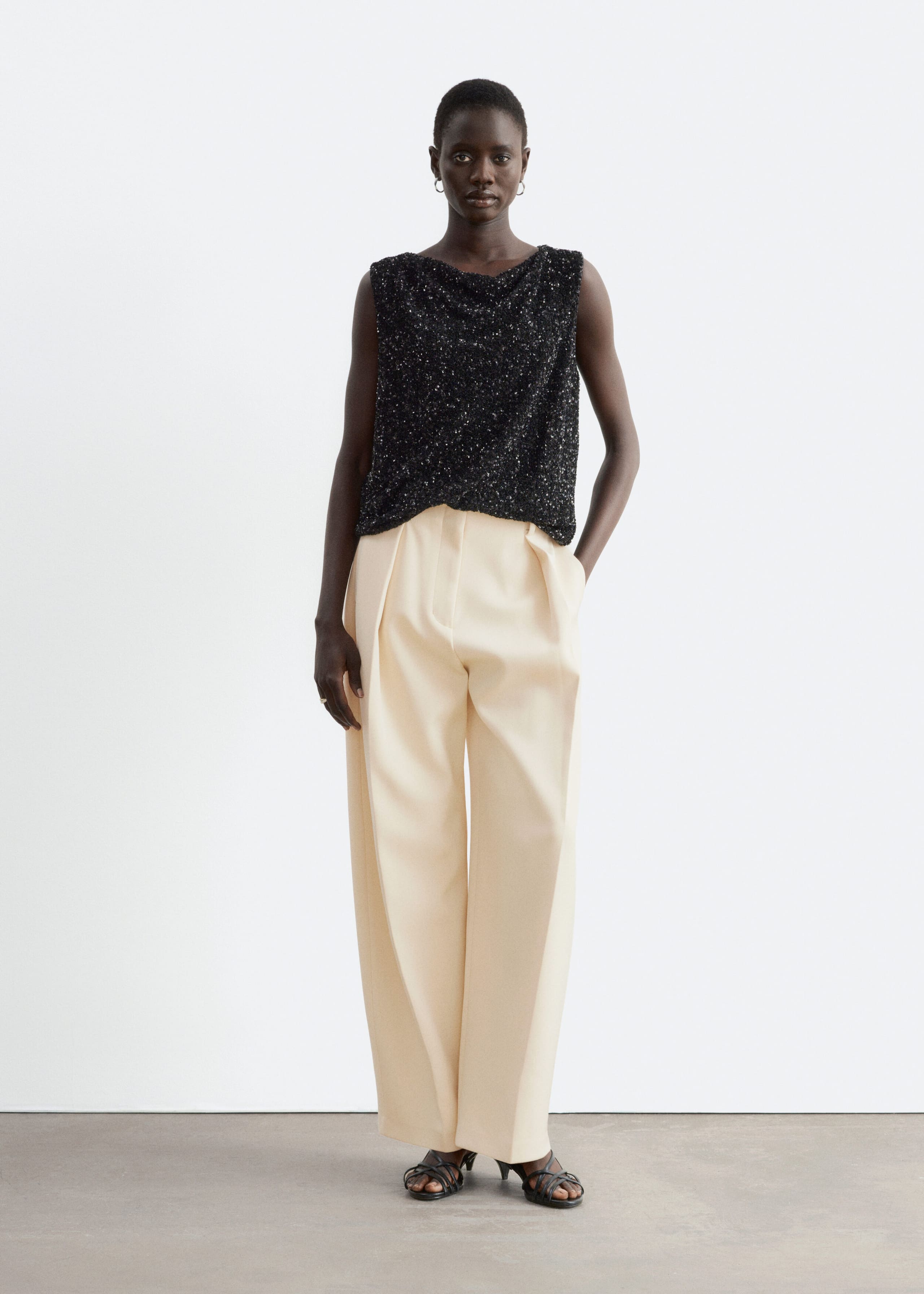 A black sleeveless mesh top covered in shimmering sequins and featuring a draped neckline is being worn by the model. The top is paired with cream-coloured, pleated wide-leg trousers and black heeled sandals.