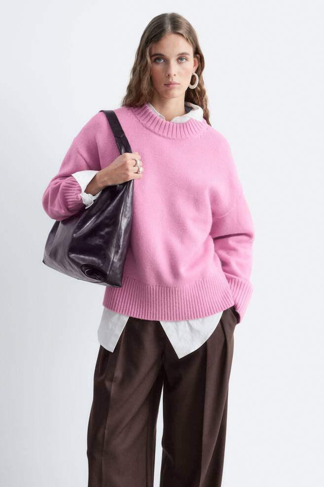 Een oversized roze wollen trui met een geribde ronde hals, verlaagde schouders en een brede geribde zoom wordt gedragen door het model. De trui is gelaagd over een wit overhemd met kraag en gecombineerd met een bruine broek, terwijl een zwarte leren schoudertas over Ã©Ã©n schouder wordt gedragen. - 1