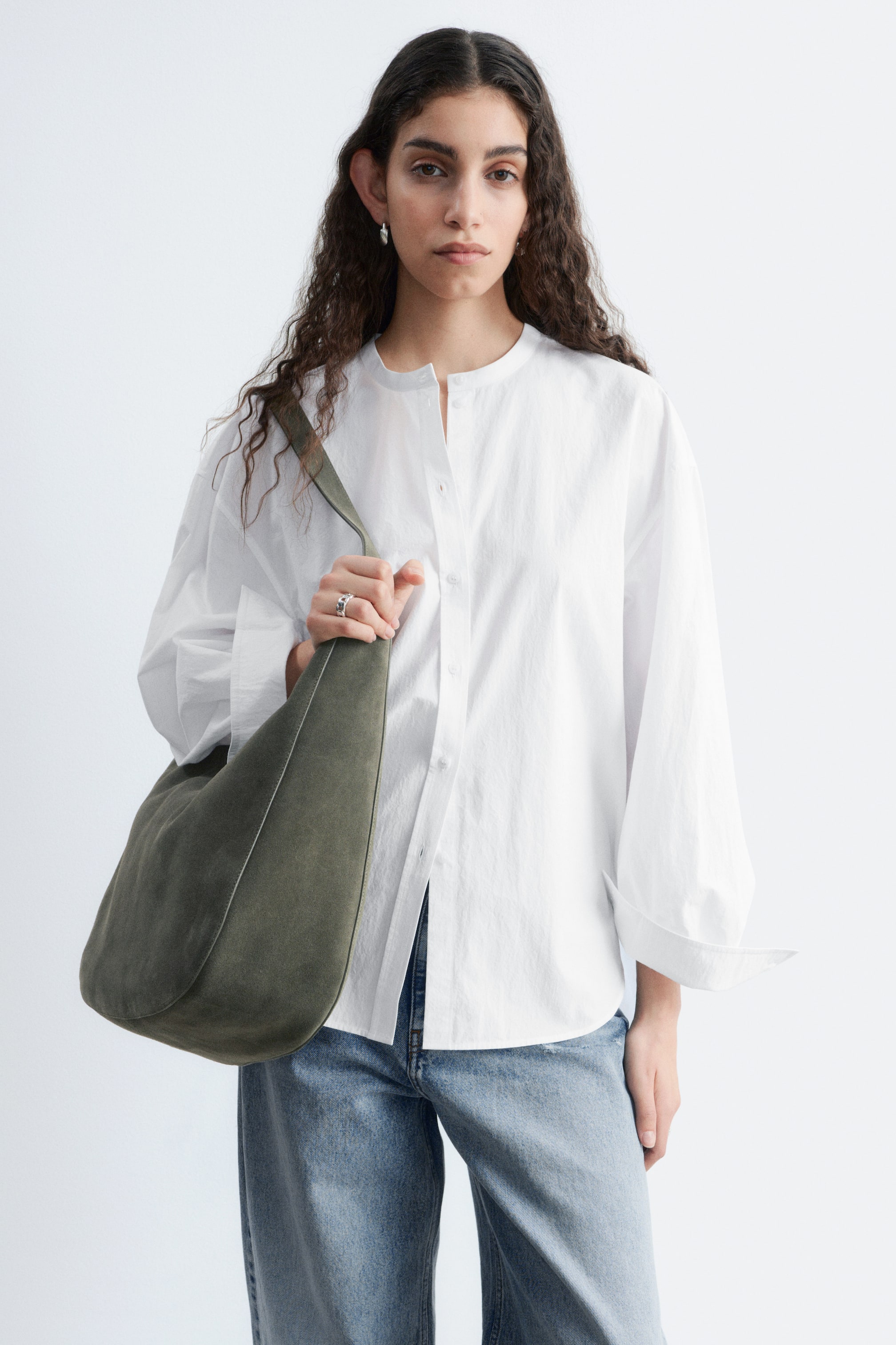 View larger image: An olive green suede shoulder bag is being worn over the left shoulder, resting against the wearer's hip. Its wide strap is visible, and a lighter-toned edge detail outlines its curved shape.