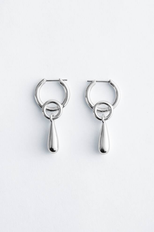 A pair of polished silver-toned hoop earrings featuring a smaller ring, from which a smooth, polished teardrop-shaped dangle is hanging. - 1