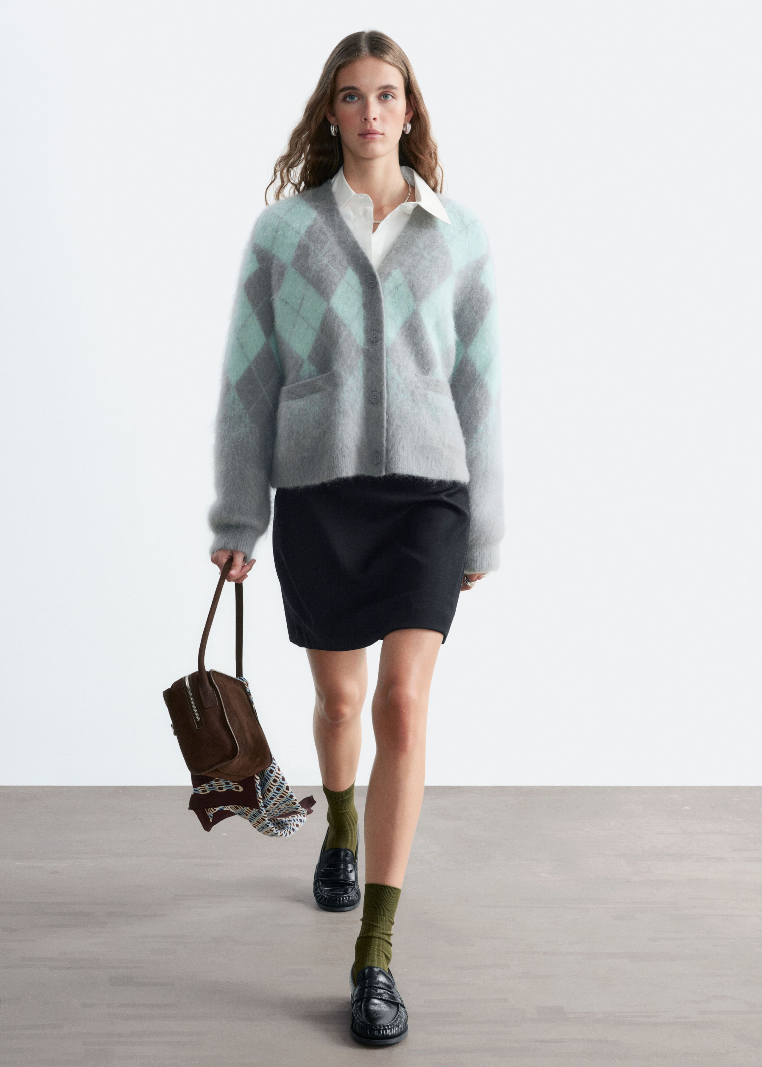 A mohair-blend argyle cardigan in gray and mint green is worn by the model. The fuzzy cardigan features a V-neck, front button closure, and patch pockets, and is layered over a white collared shirt.