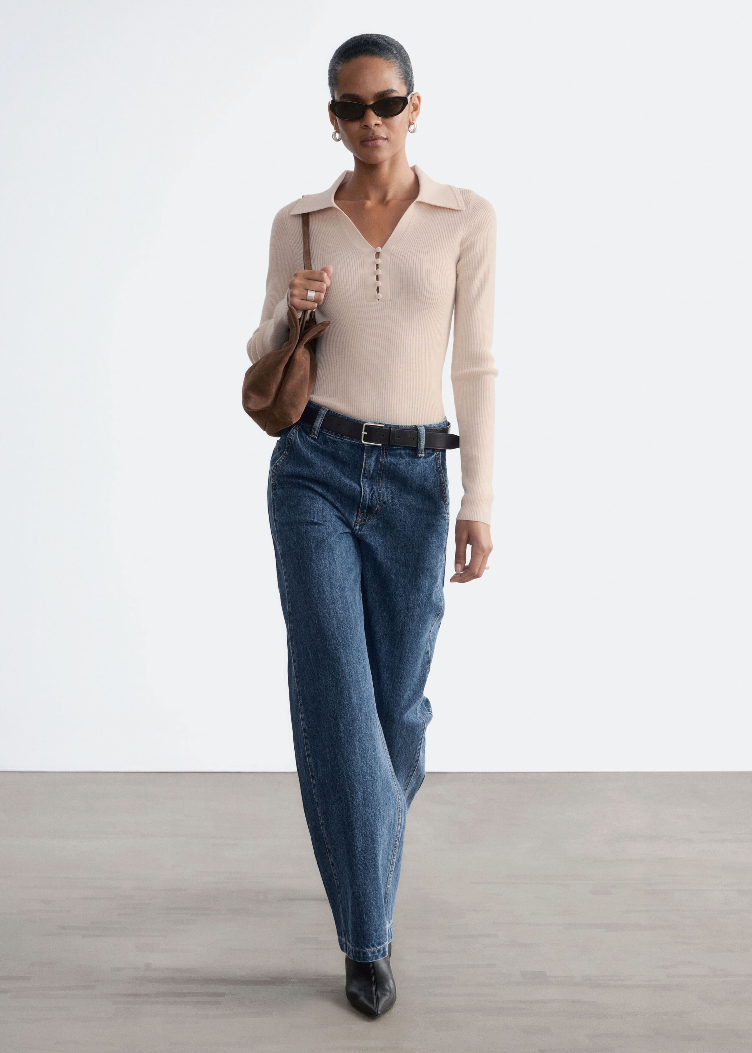 High-waisted, wide-leg blue denim jeans with slanted front pockets are worn by the model as she walks forward. The jeans are styled with a tucked-in beige long-sleeve polo knit top and a dark leather belt.