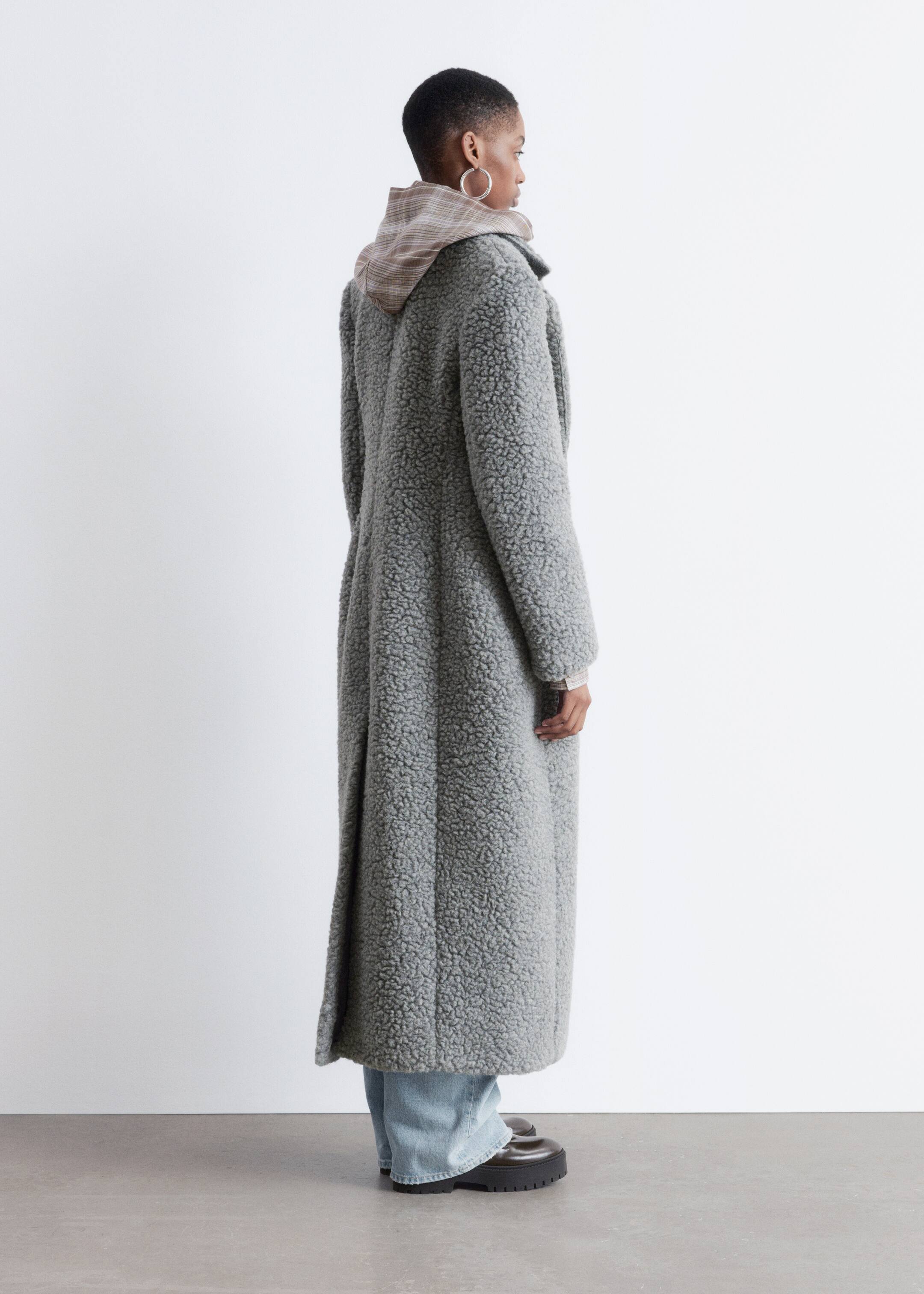View larger image: A maxi-length grey bouclé coat is being shown from a side-back angle, featuring a visible collar and a plaid hoodie layering underneath with its hood peeking over the coat's collar. Light wash jeans and dark chunky-soled shoes are visible below the coat's hem.