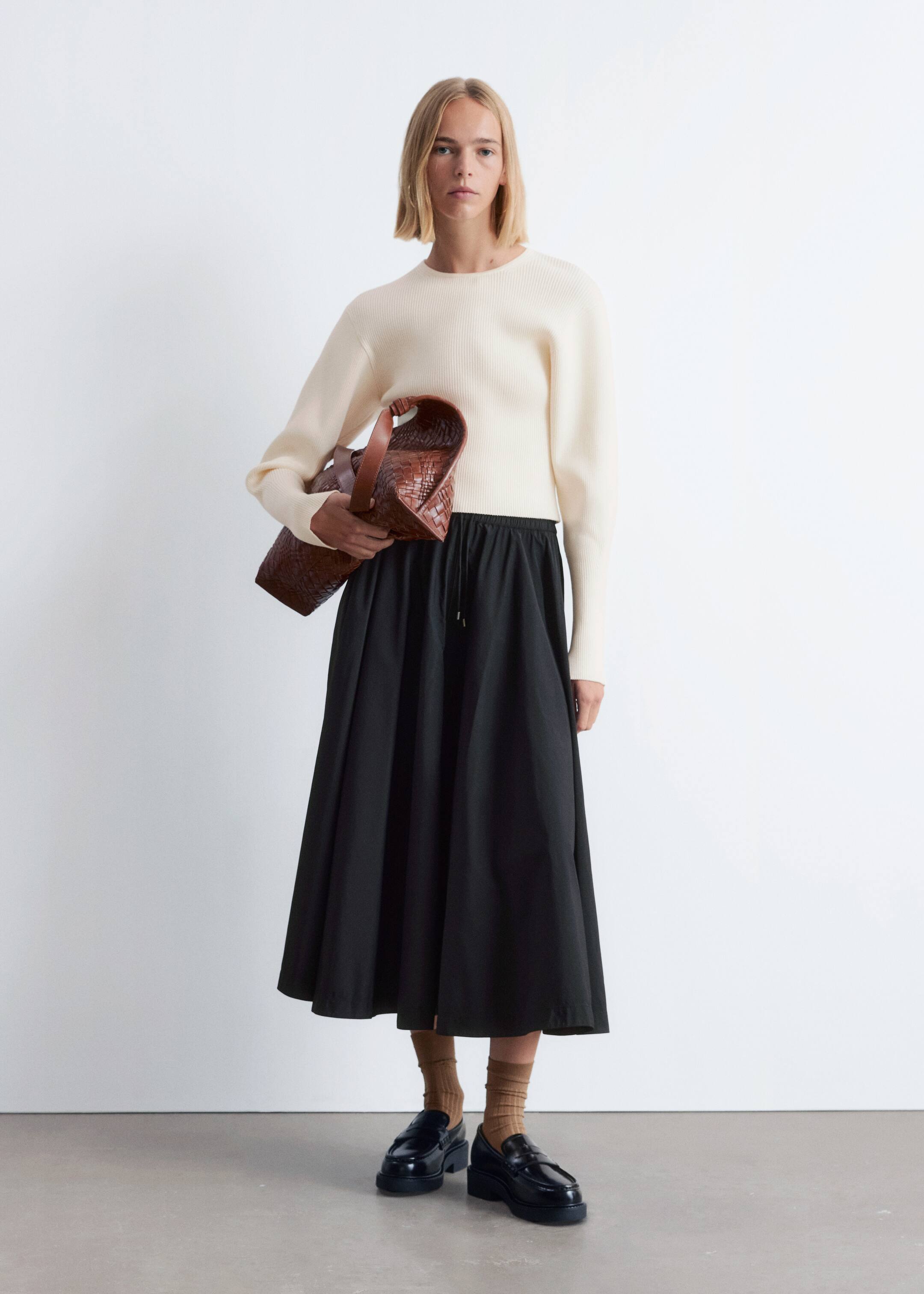 Größeres Bild anzeigen: Ein cremefarbener gerippter Strickpullover wird mit einem schwarzen Midirock getragen, der über einen elastischen Kordelzug in der Taille und eine volle, geraffte Silhouette verfügt. Eine braune geflochtene Ledertasche wird getragen, und schwarze klobige Loafer werden mit hellbraunen strukturierten Socken getragen.