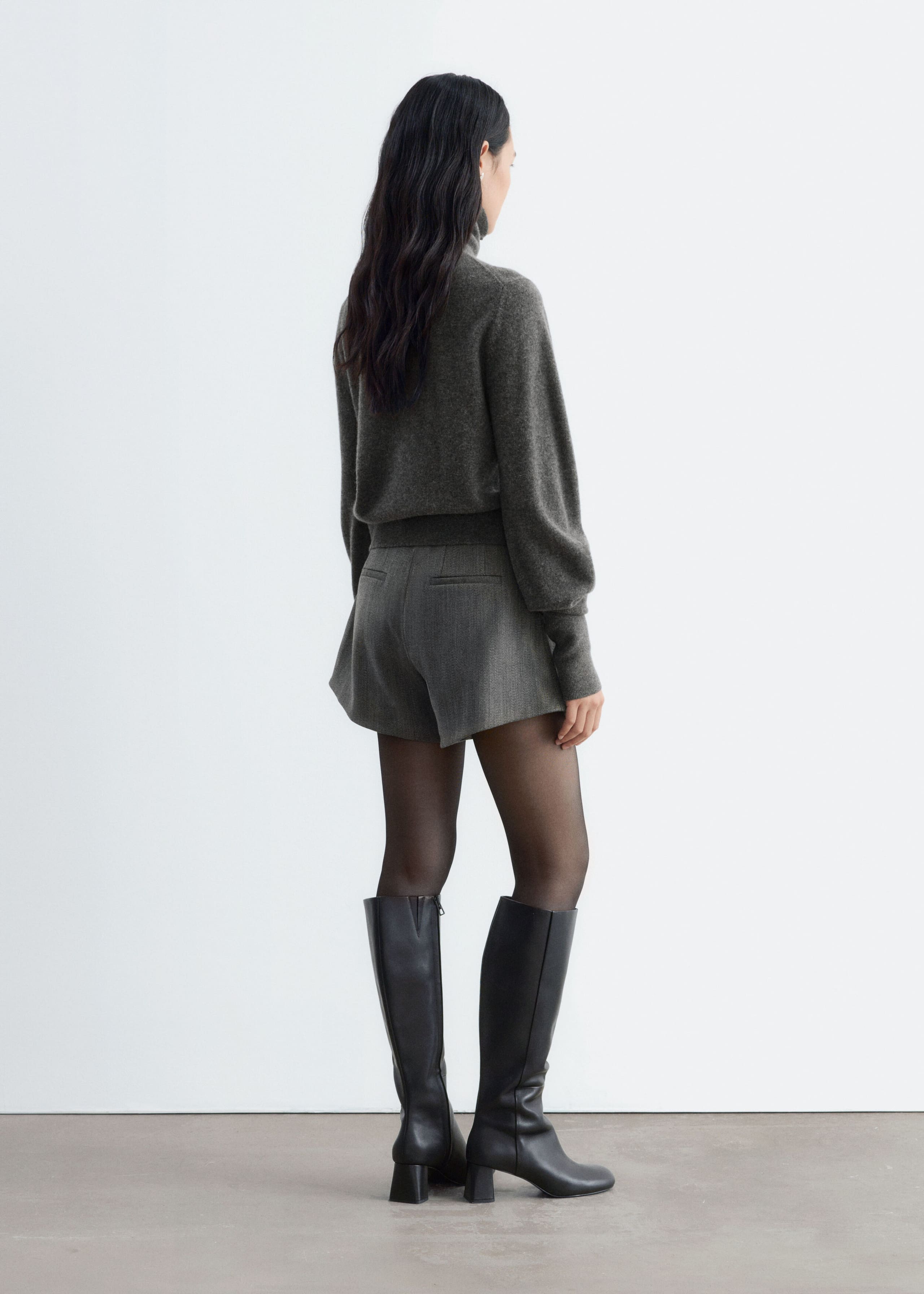 Grey, high-waisted wool-blend shorts featuring a flared A-line silhouette and back welt pockets are worn by the model. The shorts are styled with a dark grey mock-neck sweater, sheer black tights, and black knee-high leather boots.