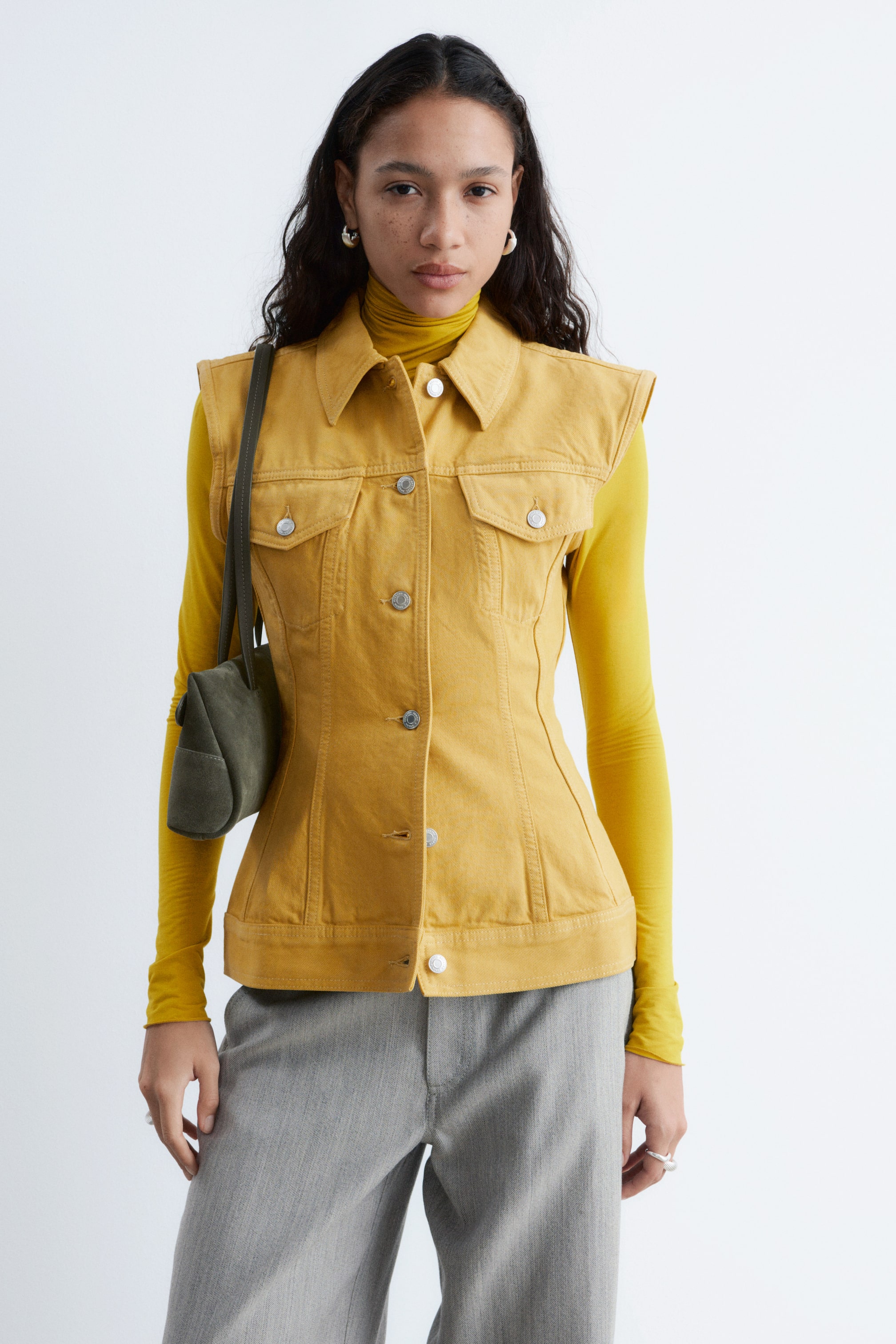 Ver imagen más grande: Un chaleco de mezclilla amarillo presenta un cuello en punta, un cierre frontal con botones plateados y dos bolsillos con solapa en el pecho. Las costuras verticales prominentes crean una silueta ajustada, y está superpuesto sobre un jersey de cuello alto amarillo de manga larga.