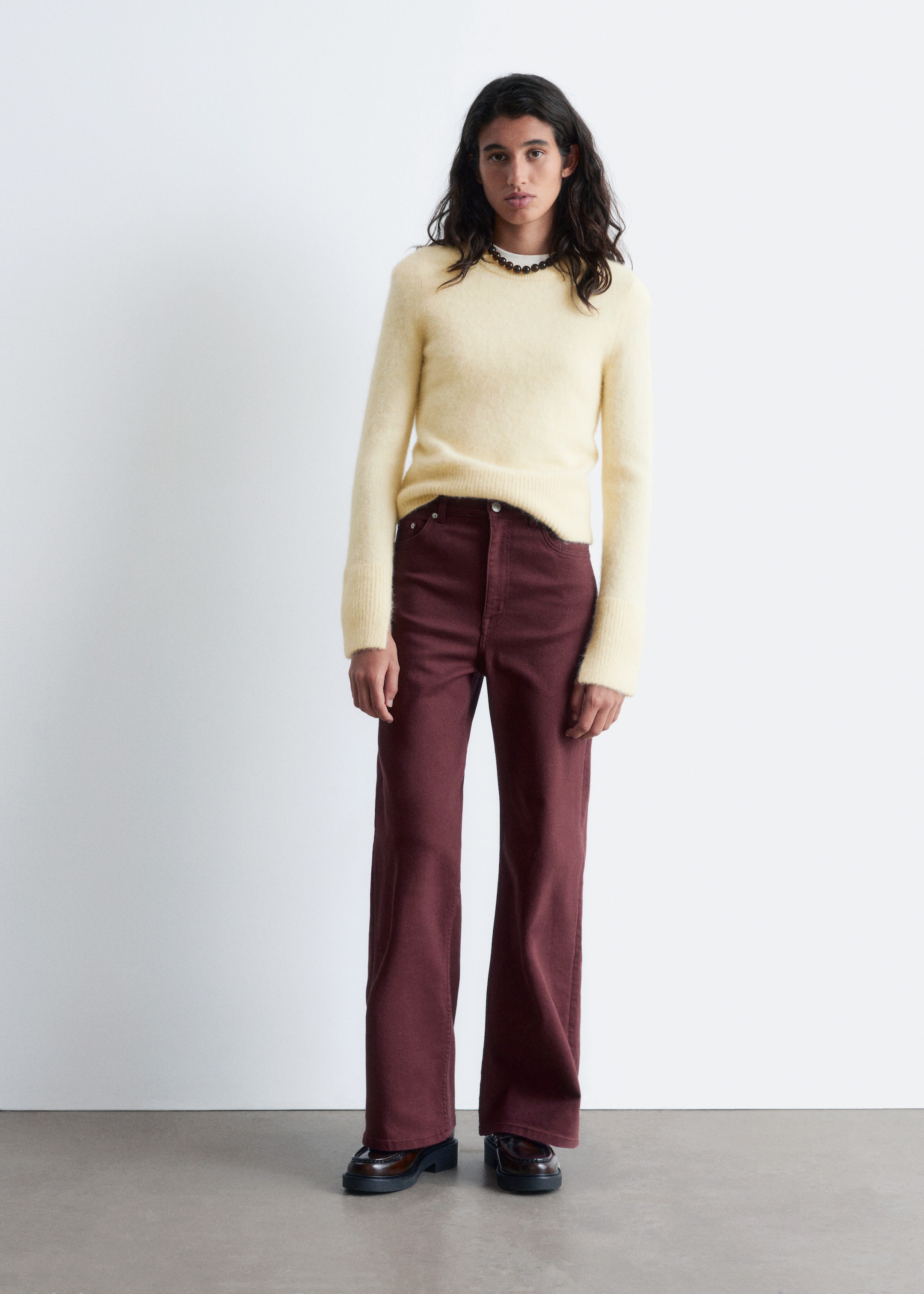 Ingrandisci l'immagine: Un maglione girocollo giallo chiaro e peloso viene indossato con pantaloni a gamba larga rosso scuro. Il maglione presenta polsini a coste ed è leggermente corto, mentre i pantaloni sono a vita alta con una vestibilità rilassata. Mocassini chunky marrone scuro e una collana girocollo con perline scure completano l'insieme.