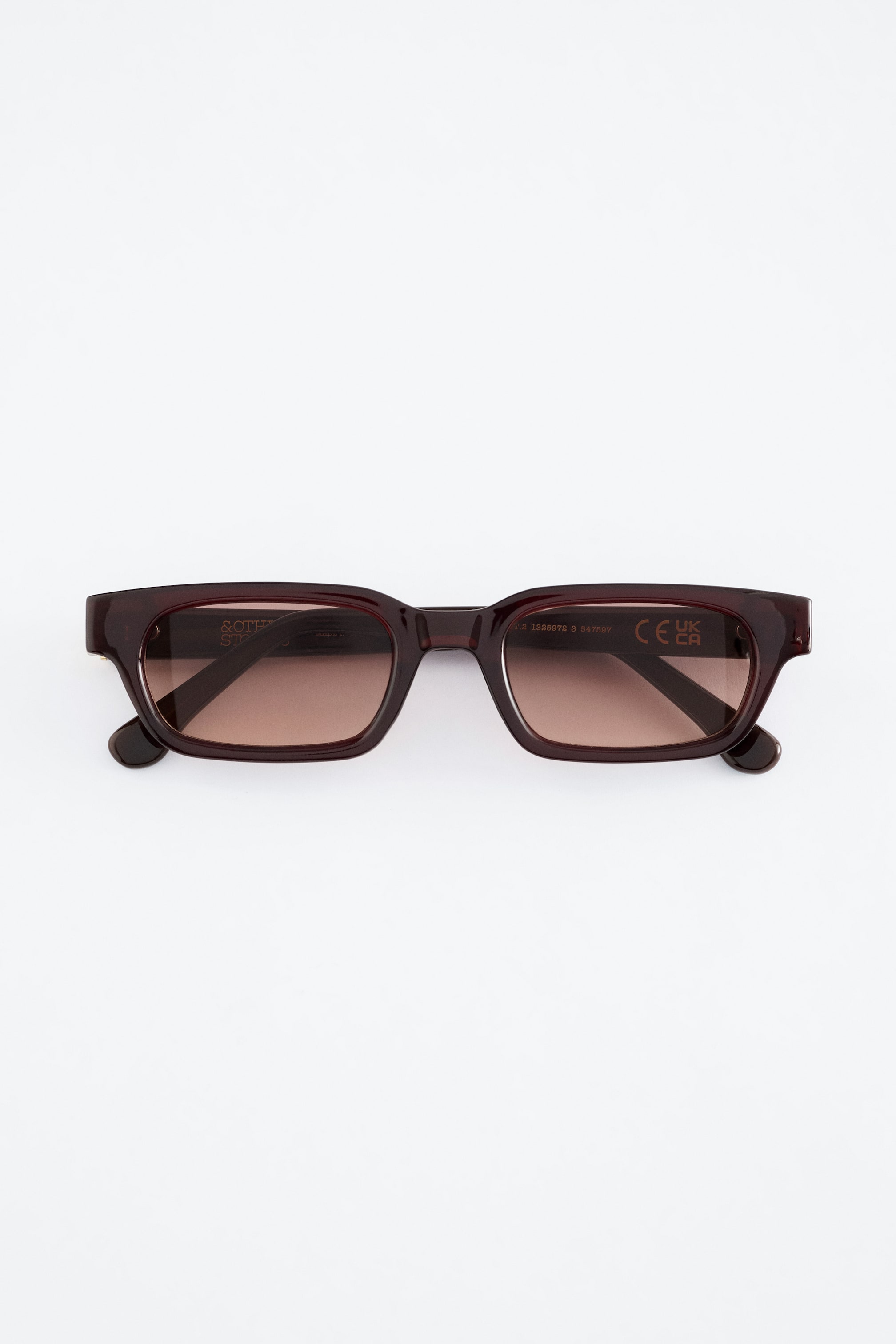 Grotere afbeelding bekijken: Smal omrande zonnebrillen tonen een rechthoekig donker roodbruin montuur met bruine gradient lenzen. De binnenzijde van de pootjes vertoont merk- en certificeringsmarkeringen.