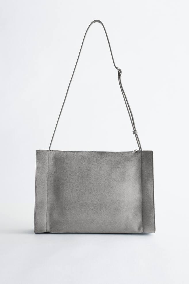 Un sac bandoulière en suède gris avec une façade rectangulaire. Il est doté d'une fine bandoulière réglable et d'une fermeture éclair le long de son bord supérieur. - 1