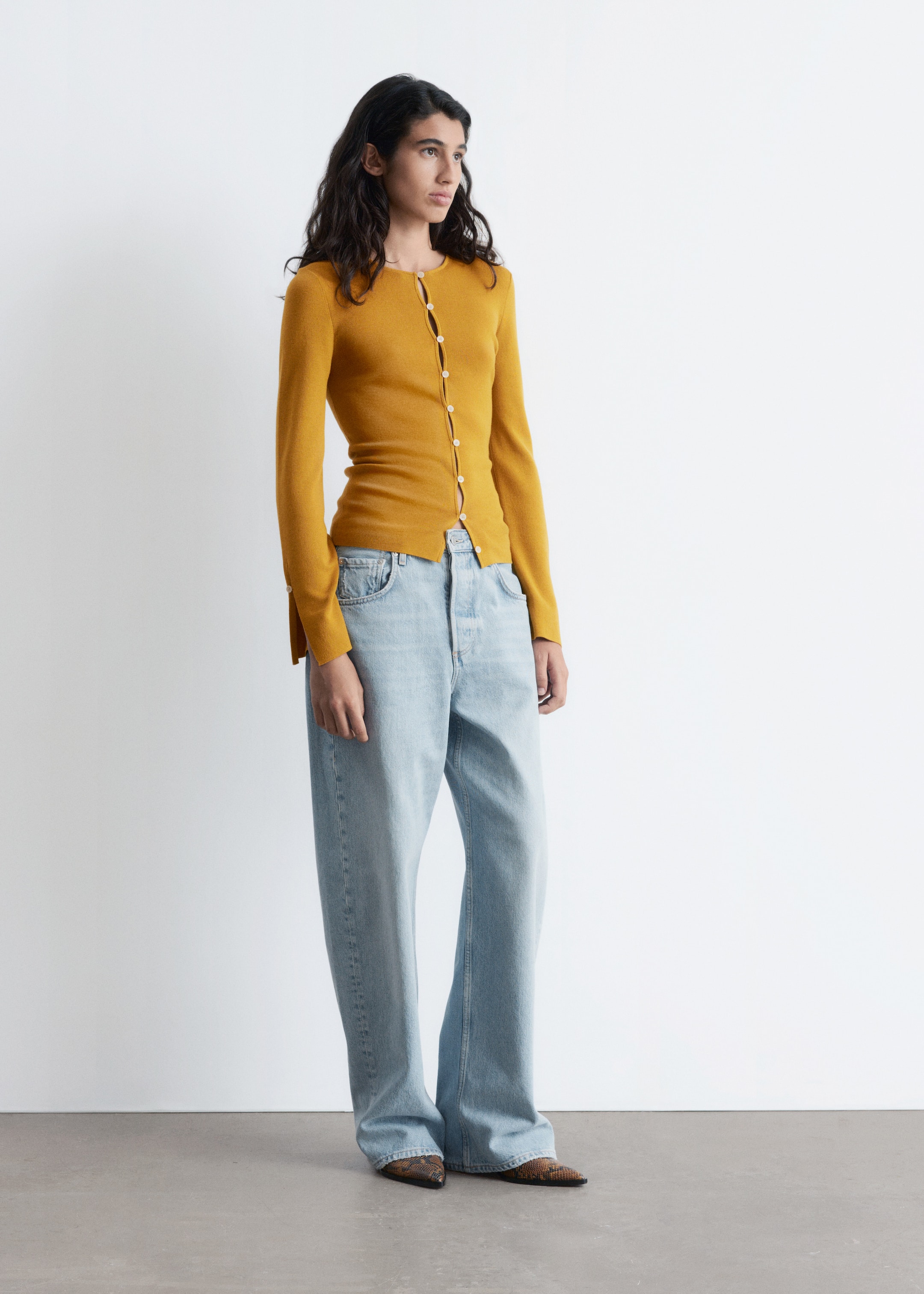 Vis større bilde: En gullfarget, slim-fit cardigan med rund hals og frontknapper bæres av modellen, som er stylet med lyse jeans og mønstrede sko. Cardiganen har lange ermer med knappede mansjetter, og dens nederste knapp er åpen, noe som skaper en utskjæringsdetalj.