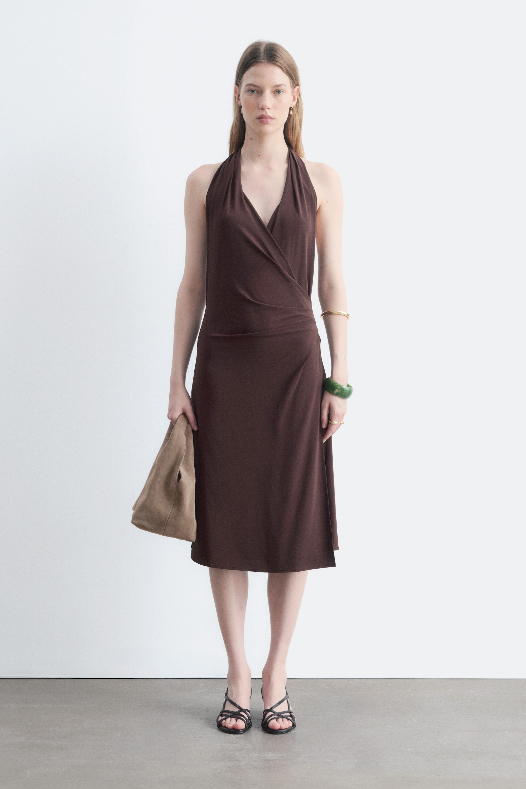 Größeres Bild ansehen: Ein braunes Wickelkleid mit Neckholder, V-Ausschnitt und drapiertem Midirock wird vom Model getragen. Dazu trägt das Model eine hellbraune Clutch und schwarze Sandalen mit Riemchen.