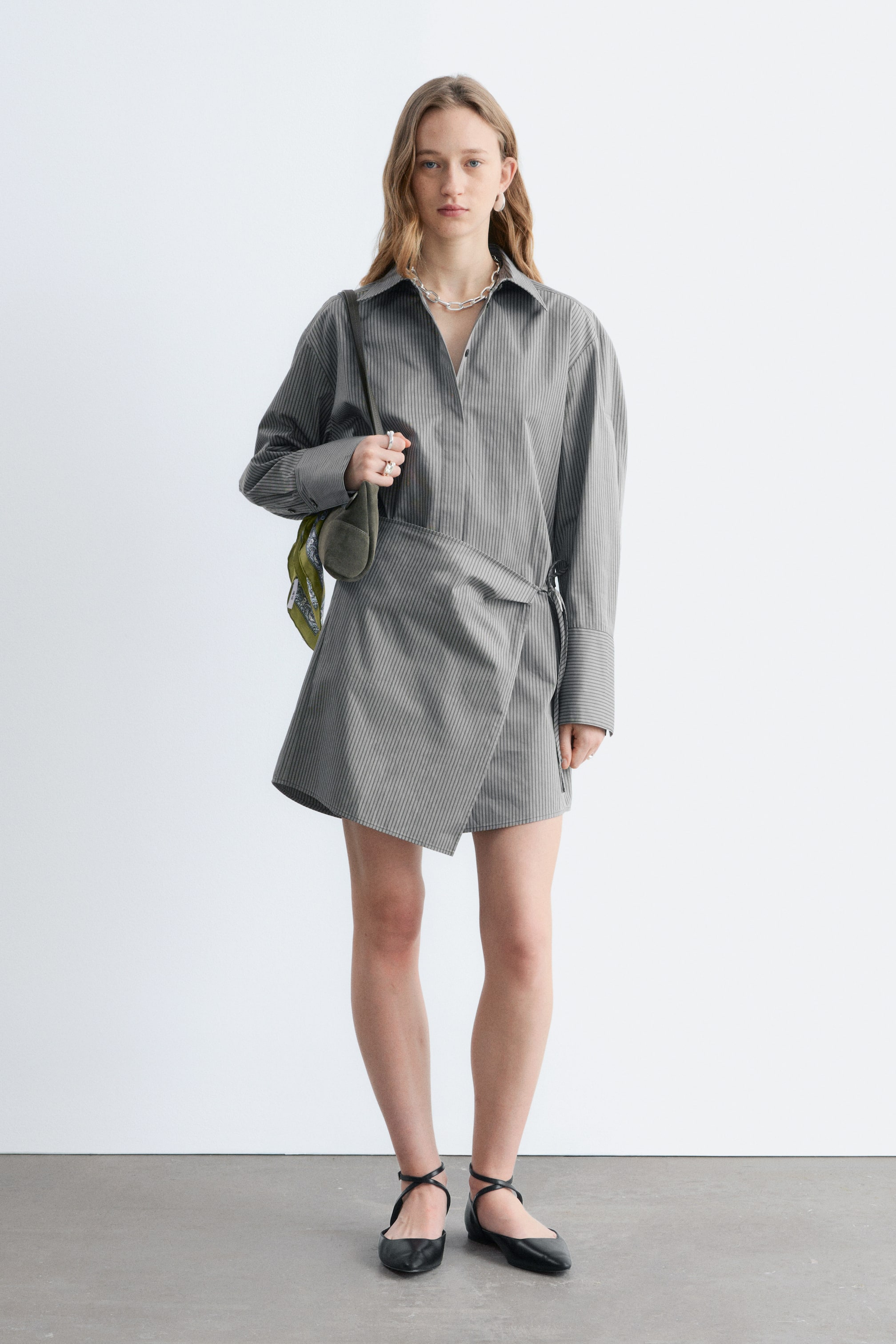 Visa större bild: En grå randig skjortklänning visas från en frontvy, med en spetsig krage, långa voluminösa ärmar med breda manschetter och en asymmetrisk omlottkjol med sidoknytning.