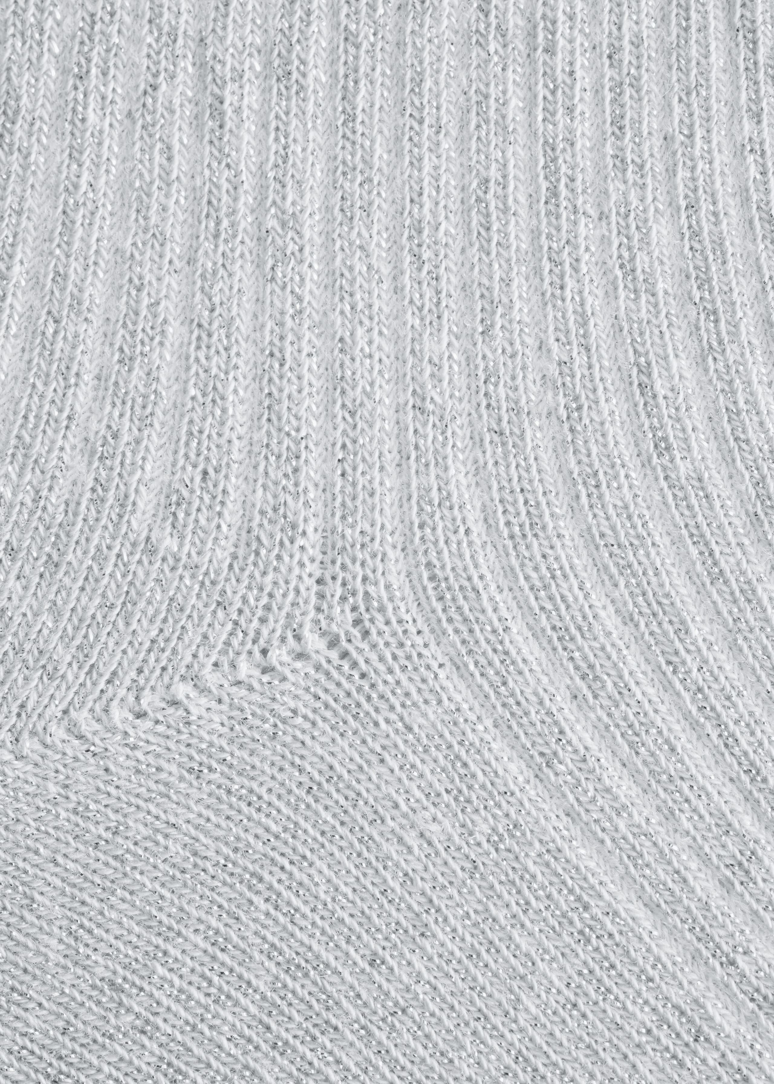 A close-up view of a light gray ribbed sock showing the detailed knit fabric woven with shimmering silver glitter lurex threads. The vertical ribbing transitions into a diagonal knit pattern, highlighting the textured metallic finish of the material.