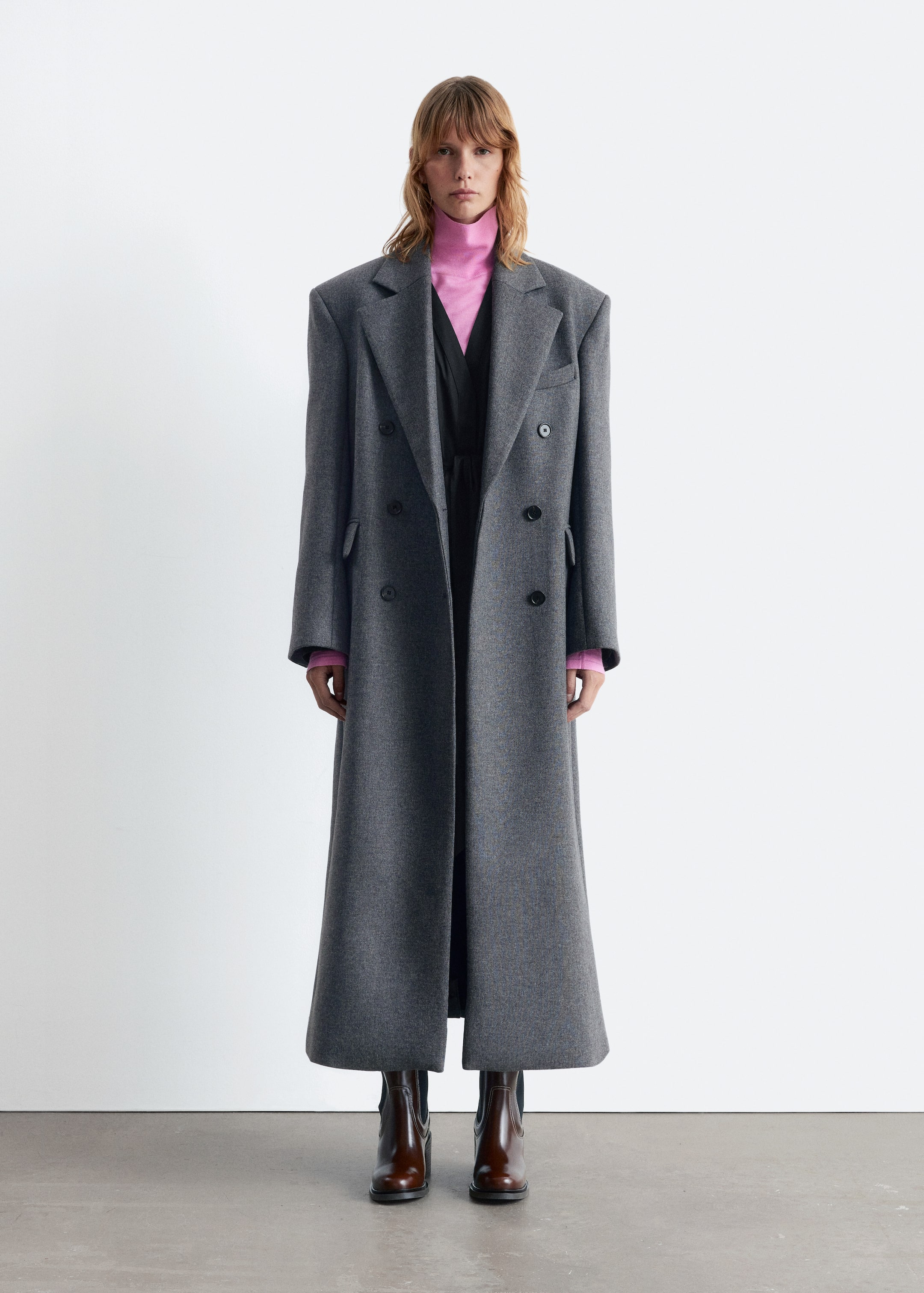 Agrandir l'image: Un long manteau gris à double boutonnage est exposé, avec de larges revers crantés, des épaules structurées et six boutons sombres sur le devant. Le manteau présente une coupe oversize avec un ourlet évasé, superposé à un col roulé rose et un vêtement noir, et s'étend jusqu'aux chevilles au-dessus de bottes marron.