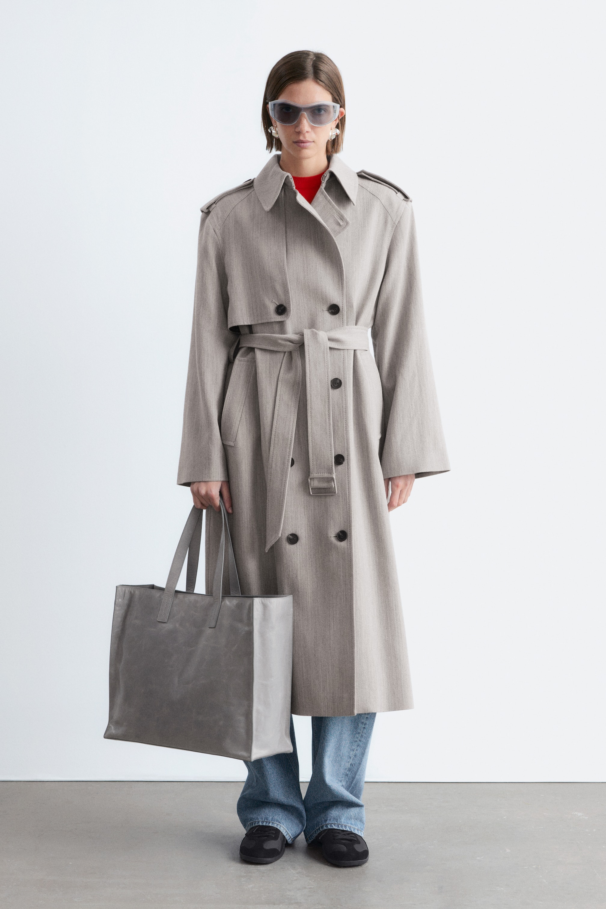 Ver imagen más grande: Un abrigo trench de doble botonadura en color greige claro con una sutil textura de espiga se muestra de frente, presentando un cuello en punta, solapas de muesca, charreteras y una solapa de tormenta en el pecho izquierdo. El abrigo se ajusta a la cintura con un cinturón de la misma tela y se extiende hasta media pantorrilla.