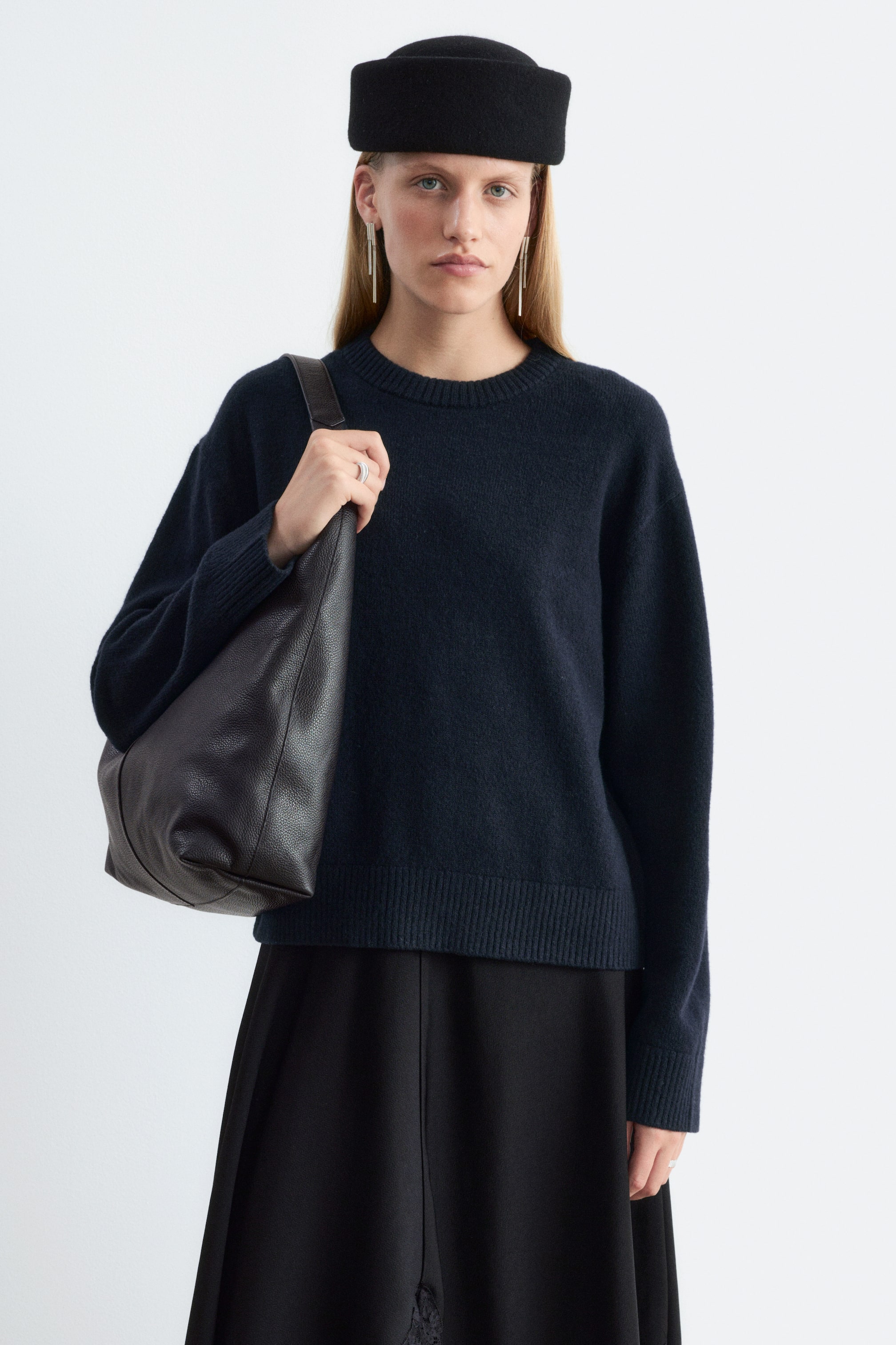 Visualizza l'immagine ingrandita: Un maglione in maglia dal taglio squadrato, in blu navy, indossato dalla modella, con scollo tondo, maniche lunghe con polsini a coste e orlo a coste. Il maglione è abbinato a una gonna nera, un cappello strutturato nero e una borsa a tracolla in pelle scura.