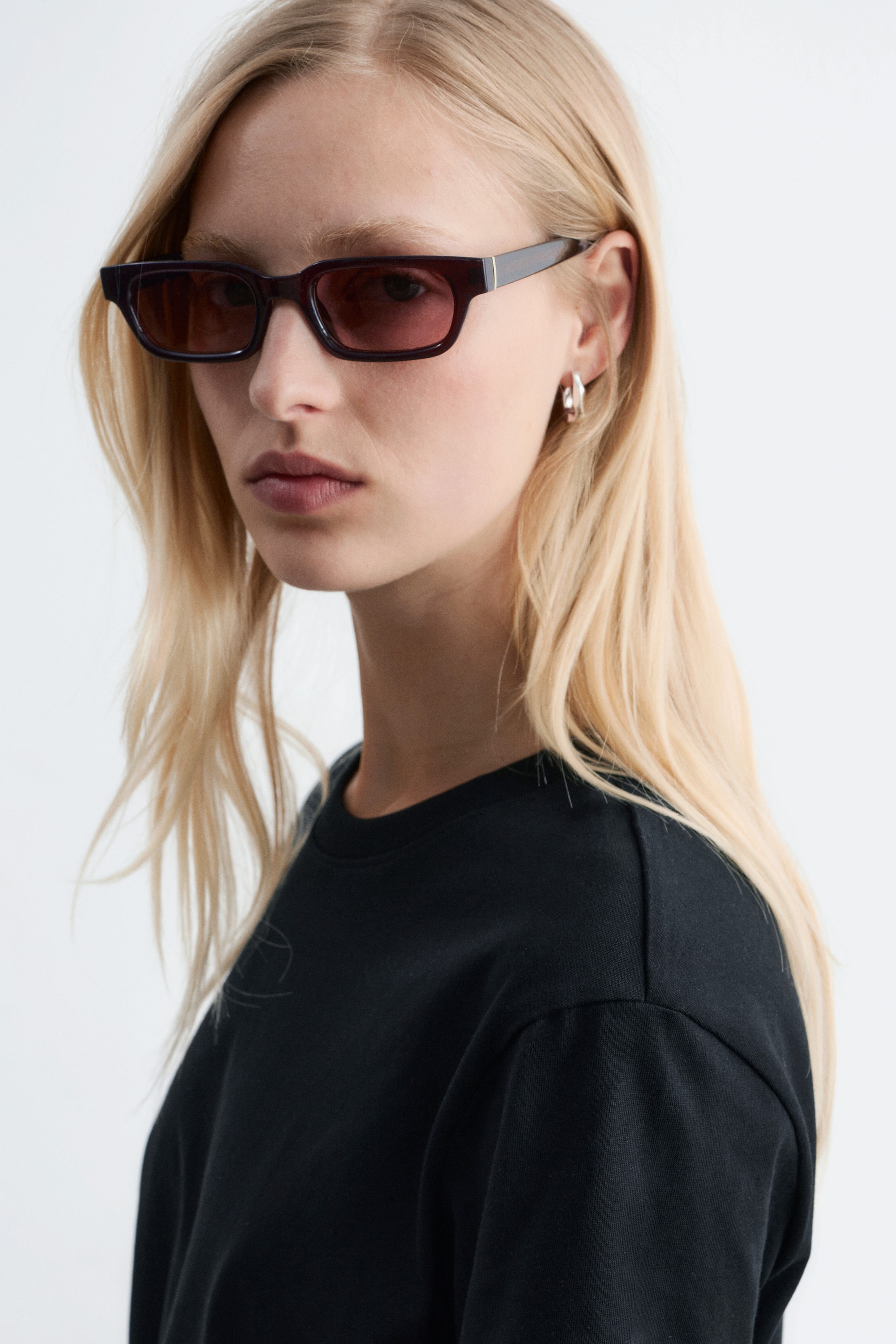 Größeres Bild anzeigen: Der/die/das Modell trägt schmal gerahmte Sonnenbrillen mit dunklen, rechteckigen Fassungen, rötlich-braun getönten Gläsern und einem dezenten goldenen Akzent am Bügel. Außerdem trägt der/die/das Modell ein schwarzes T-Shirt mit Rundhalsausschnitt.