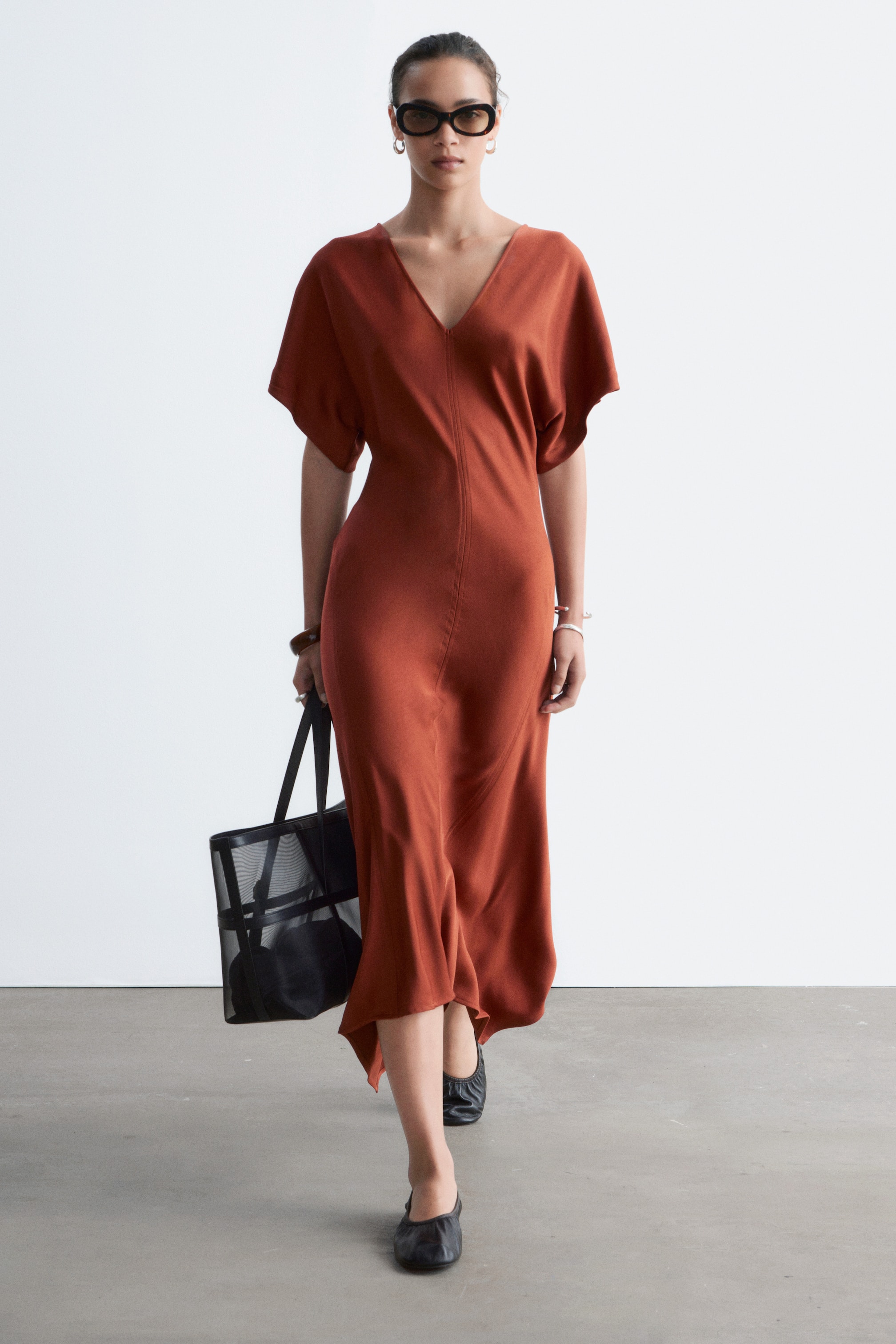 Agrandir l'image: Une robe midi couleur rouille présente un décolleté en V et des manches courtes et larges, avec une couture verticale qui descend le long de l'avant. La robe tombe à une longueur midi, affichant une silhouette fluide.