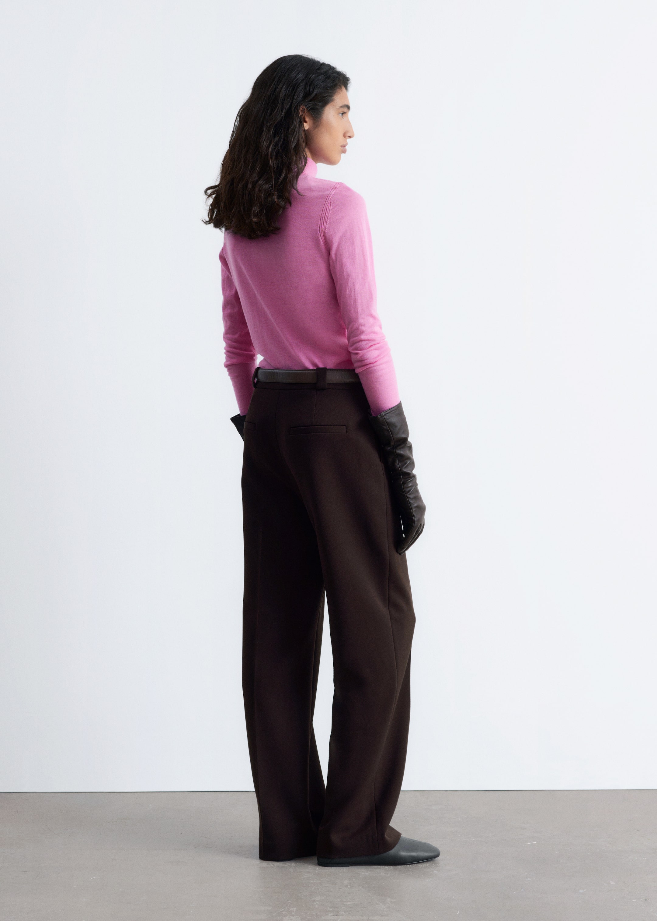 Agrandir l'image: Un pull à col roulé rose et un pantalon large marron foncé sont présentés, vus de l'arrière-droit. Le pull à col roulé rose à manches longues est porté rentré dans le pantalon large marron foncé à taille haute. Le pantalon est stylé avec une ceinture foncée visible et présente une seule poche passepoilée à l'arrière.