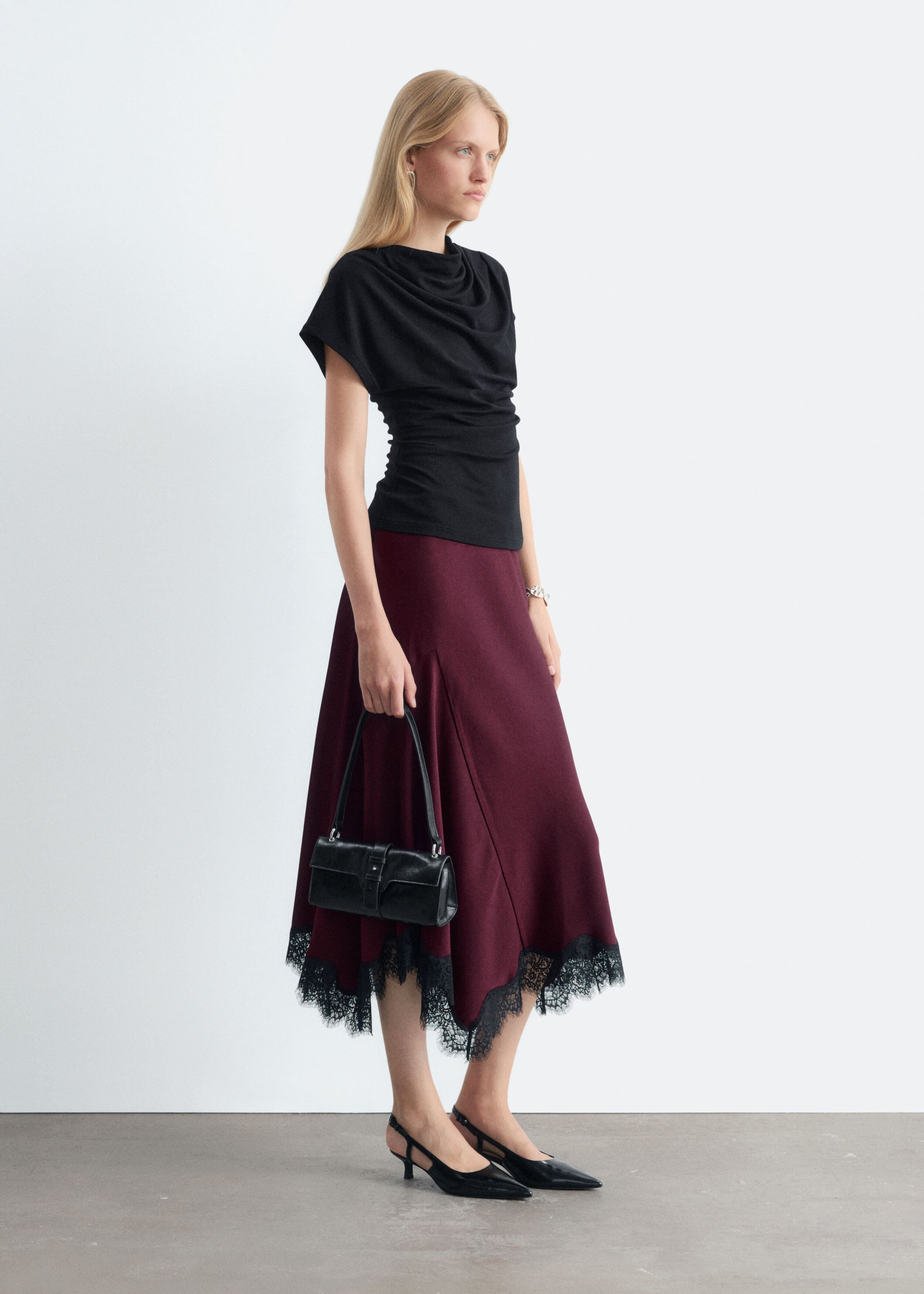 A burgundy flared midi skirt with an asymmetrical hemline and delicate black lace trim is styled on the model. The skirt is paired with a sleeveless black cowl-neck top, black slingback heels, and a small black leather handbag.