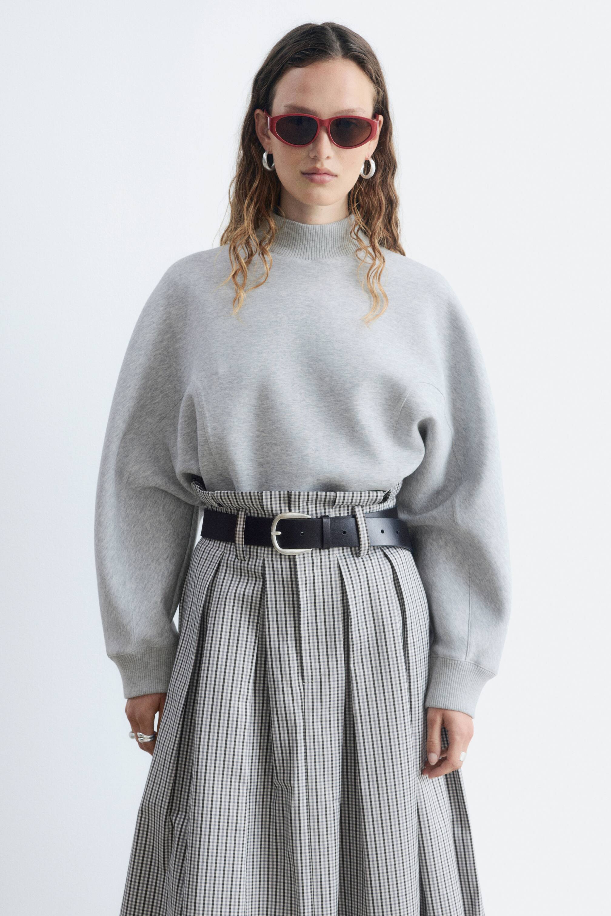 Ver imagem maior: Uma sweatshirt cinzenta de jersey com acabamento em scuba apresenta uma gola alta canelada, mangas curvas e punhos e bainha canelados. A modelo está a usar a sweatshirt, que está metida numa saia plissada e ajustada com um cinto preto na cintura.