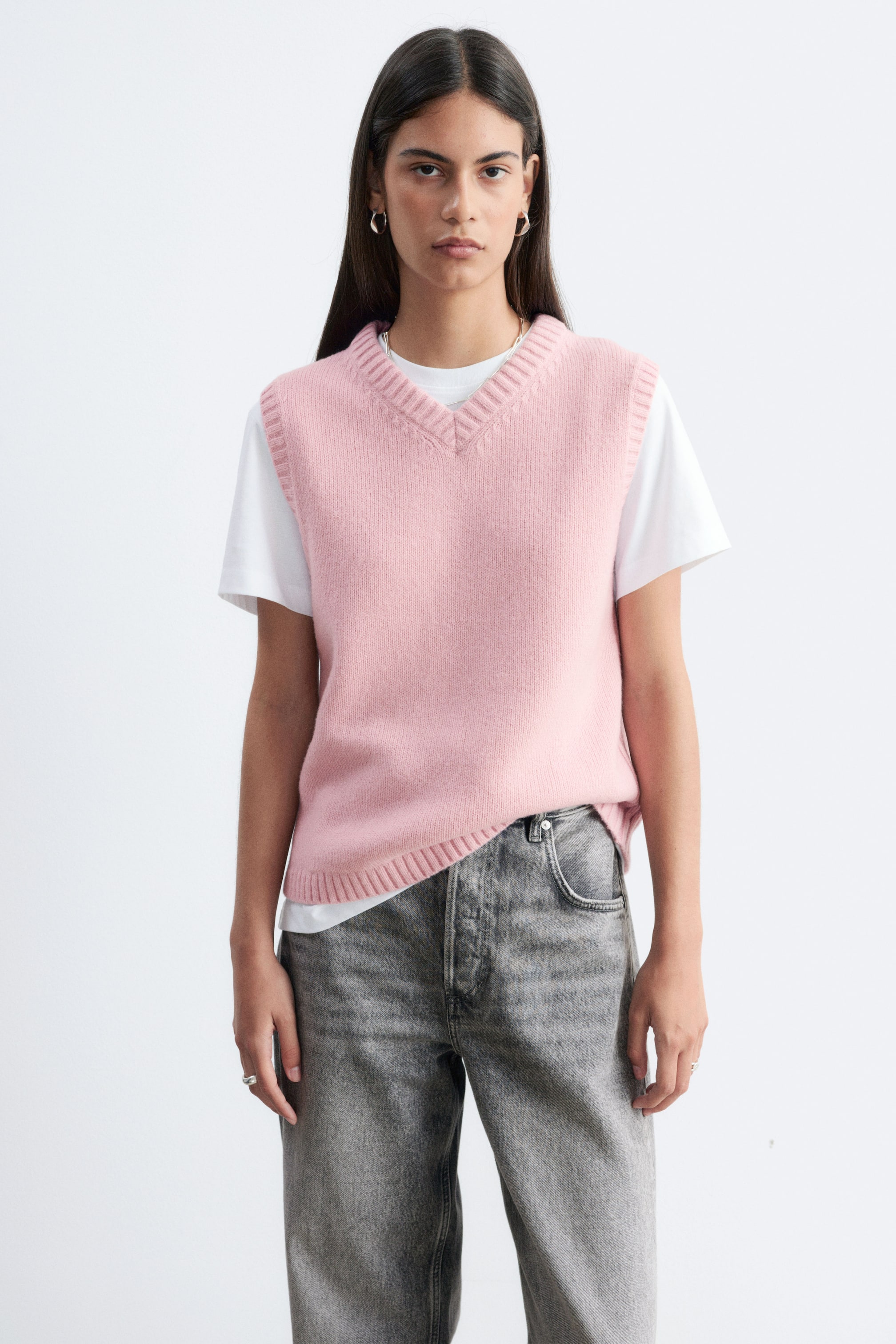 Vis større bilde: En rosa strikket vest med V-hals og ribbestrikkede kanter bæres av modellen. Vesten er lagt over en hvit kortermet T-skjorte og kombinert med grå jeans.
