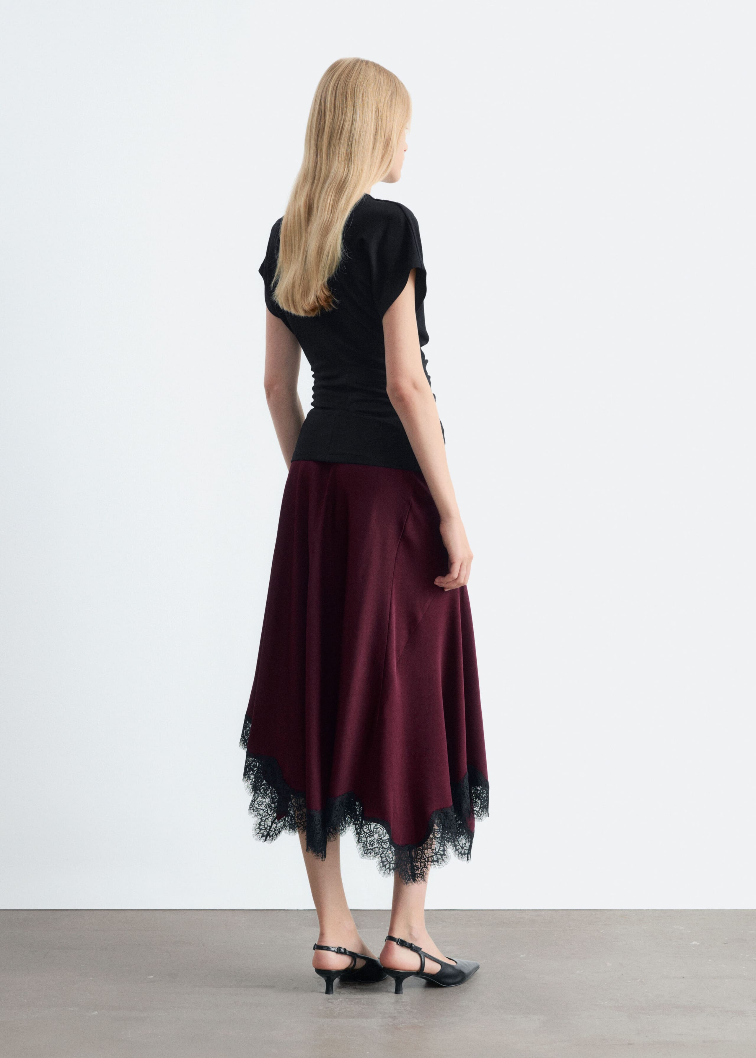The model is wearing a burgundy flared midi skirt featuring an asymmetrical hemline accented with black lace trim. The skirt is styled with a black short-sleeved top and black slingback kitten heels.