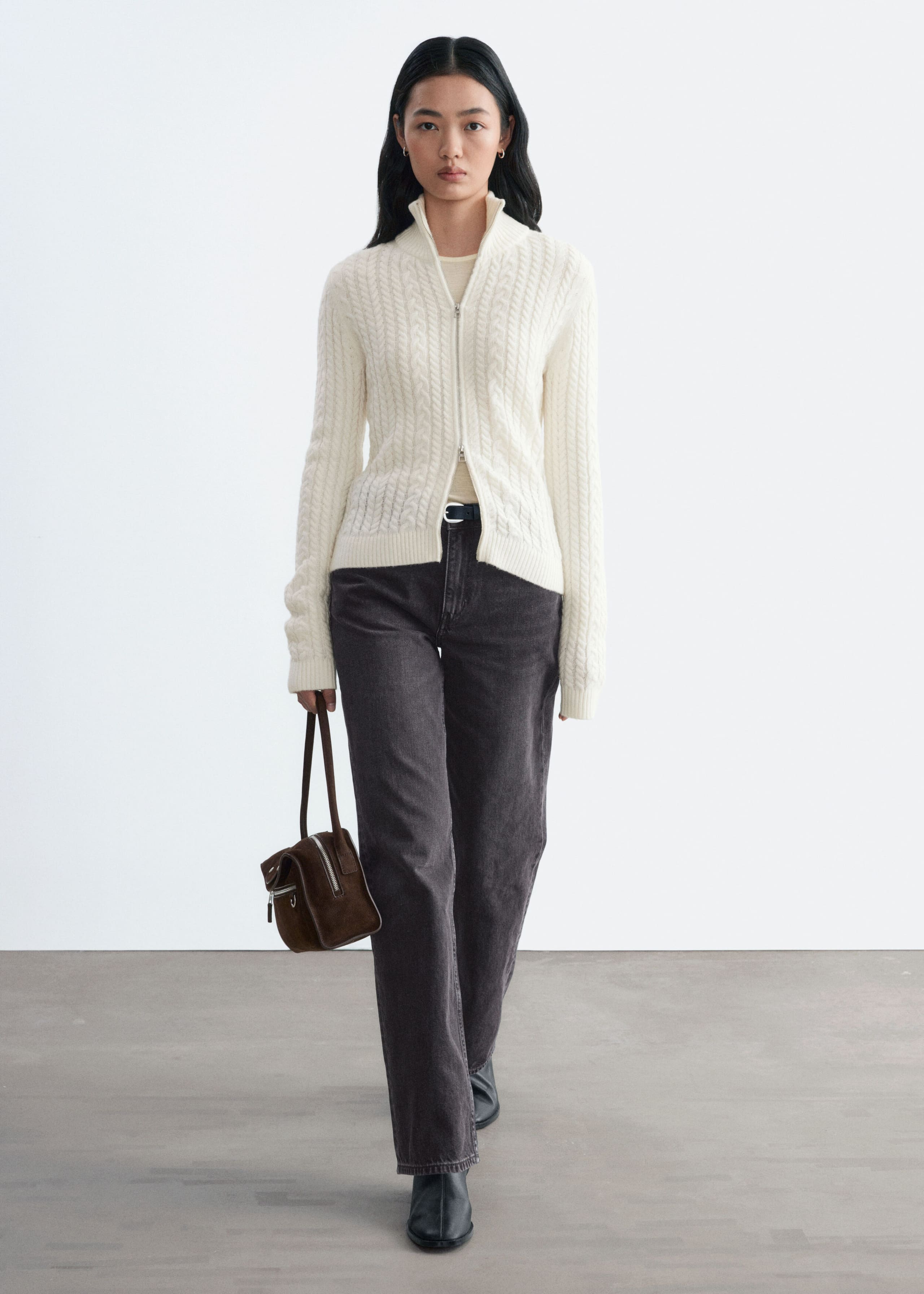 An off-white, slim-fit cardigan featuring mixed cable-knit patterns, a high ribbed collar and a two-way front zip is being worn by the model. The cardigan is styled with dark grey jeans, and the model is carrying a small brown handbag.