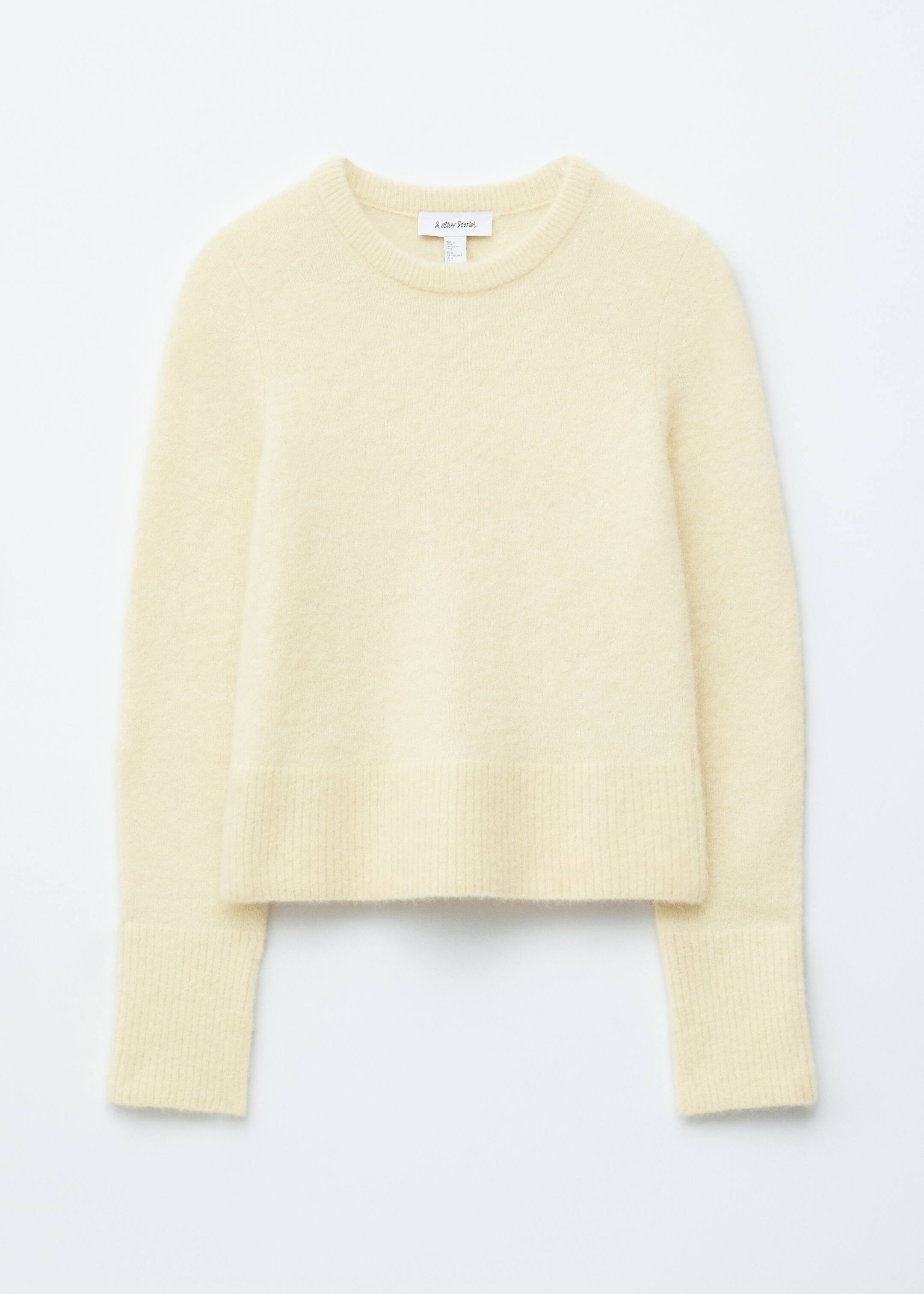 Agrandir l'image: Un pull jaune pâle, présenté de face et posé à plat, présente une texture tricotée douce et duveteuse, un col rond, des manches longues et des finitions côtelées au niveau du col, de l'ourlet et des poignets, avec des fentes subtiles sur les poignets.