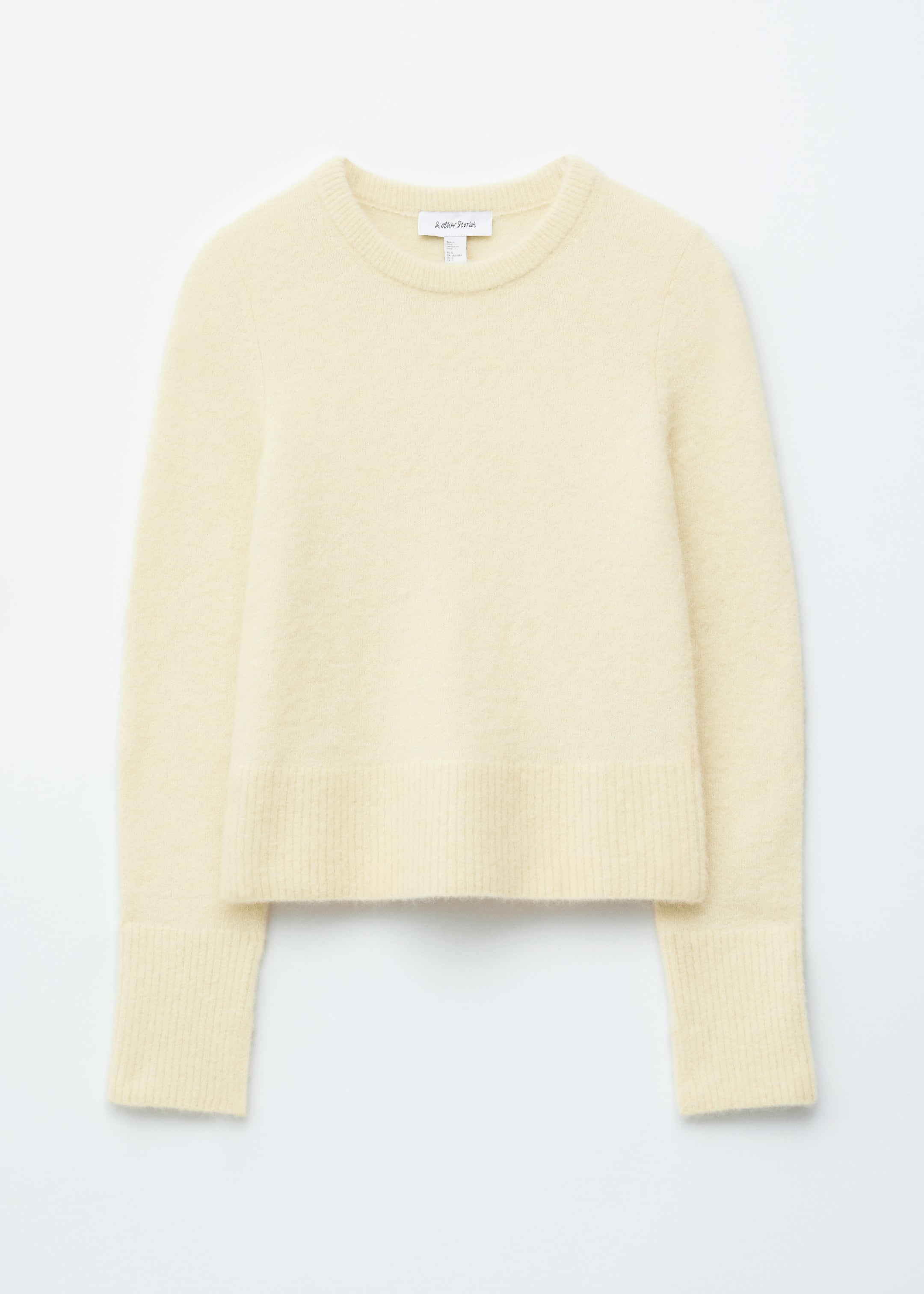 Ingrandisci l'immagine: Un maglione giallo pallido, mostrato di fronte e disteso piatto, presenta una texture morbida e pelosa, uno scollo rotondo, maniche lunghe e bordi a coste sullo scollo, sull'orlo e sui polsini, con sottili spacchi sui polsini.