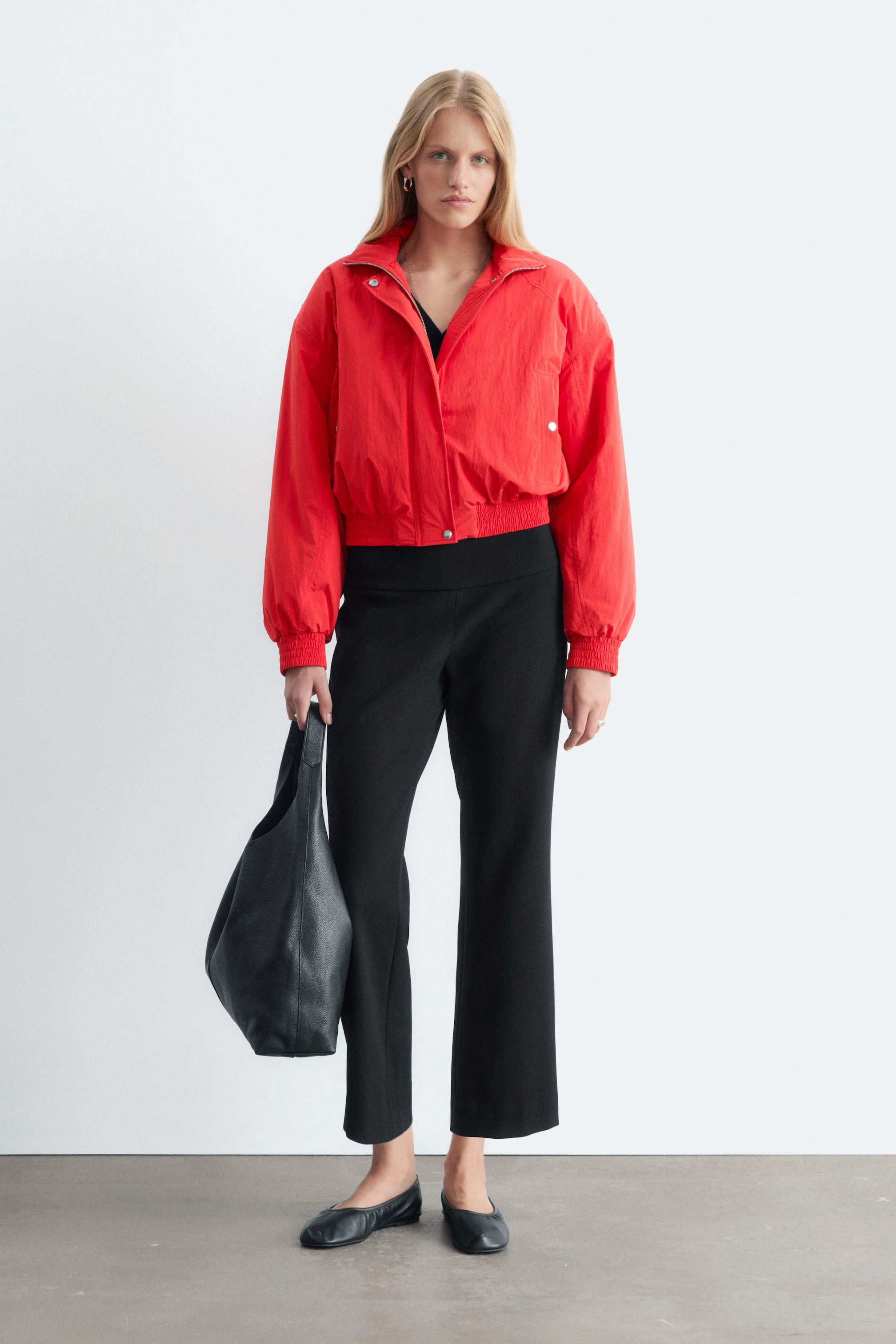 Größeres Bild anzeigen: Die in Schwarz gehaltene, cropped Kick-Flare-Hose wird vom Model getragen und zeichnet sich durch einen glatten Bund sowie eine schmale Passform aus, die am knöchellangen Saum leicht ausgestellt ist. Die Hose wird mit einer leuchtend roten Zip-up-Jacke, schwarzen Leder-Ballerinas und einer schwarzen Ledertasche kombiniert.