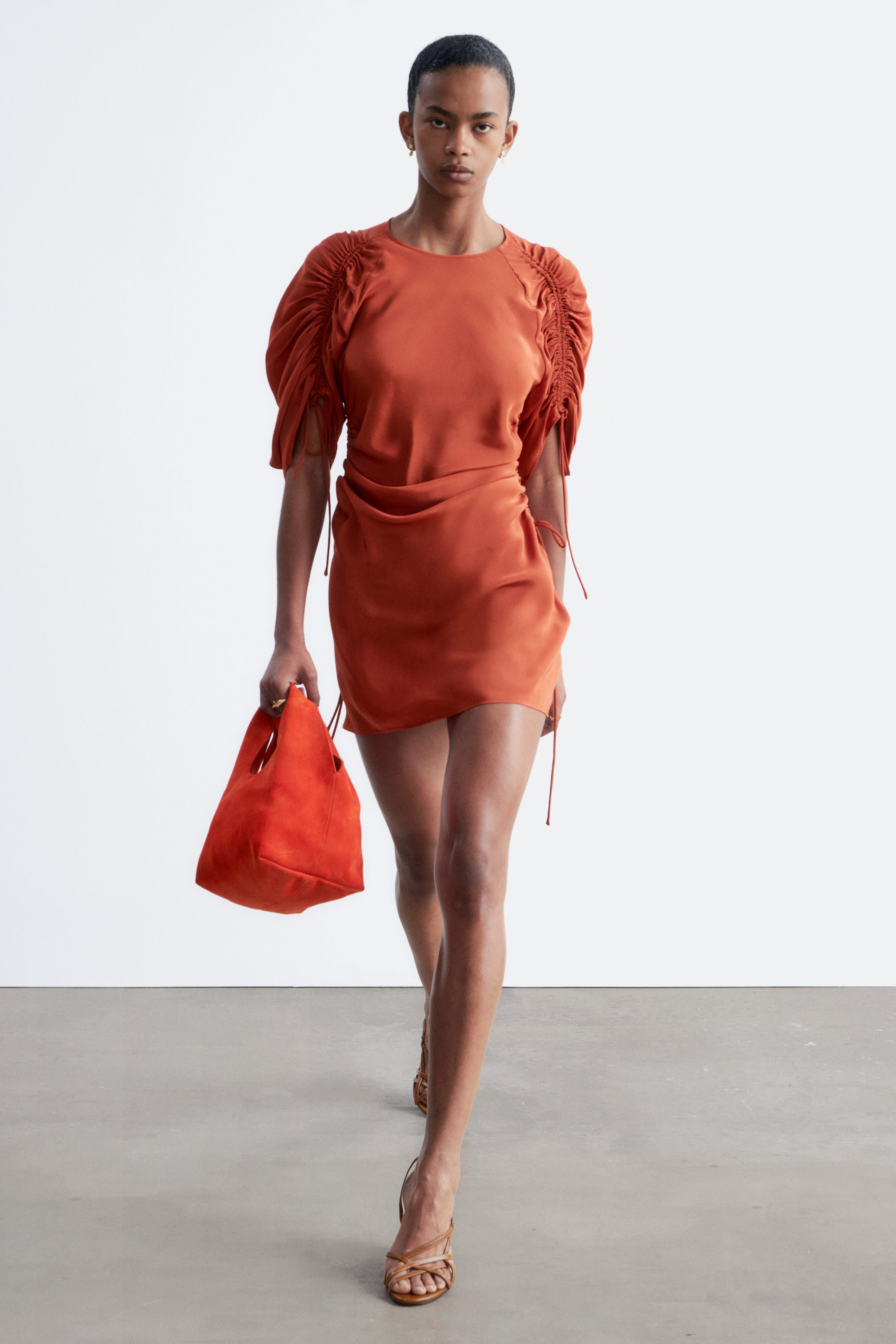 Visa större bild: En rostfärgad miniklänning med rund halsringning, voluminösa korta ärmar med rynkor och synliga knytband, samt en draperad, rynkad minikjol med knytband på ena sidan.