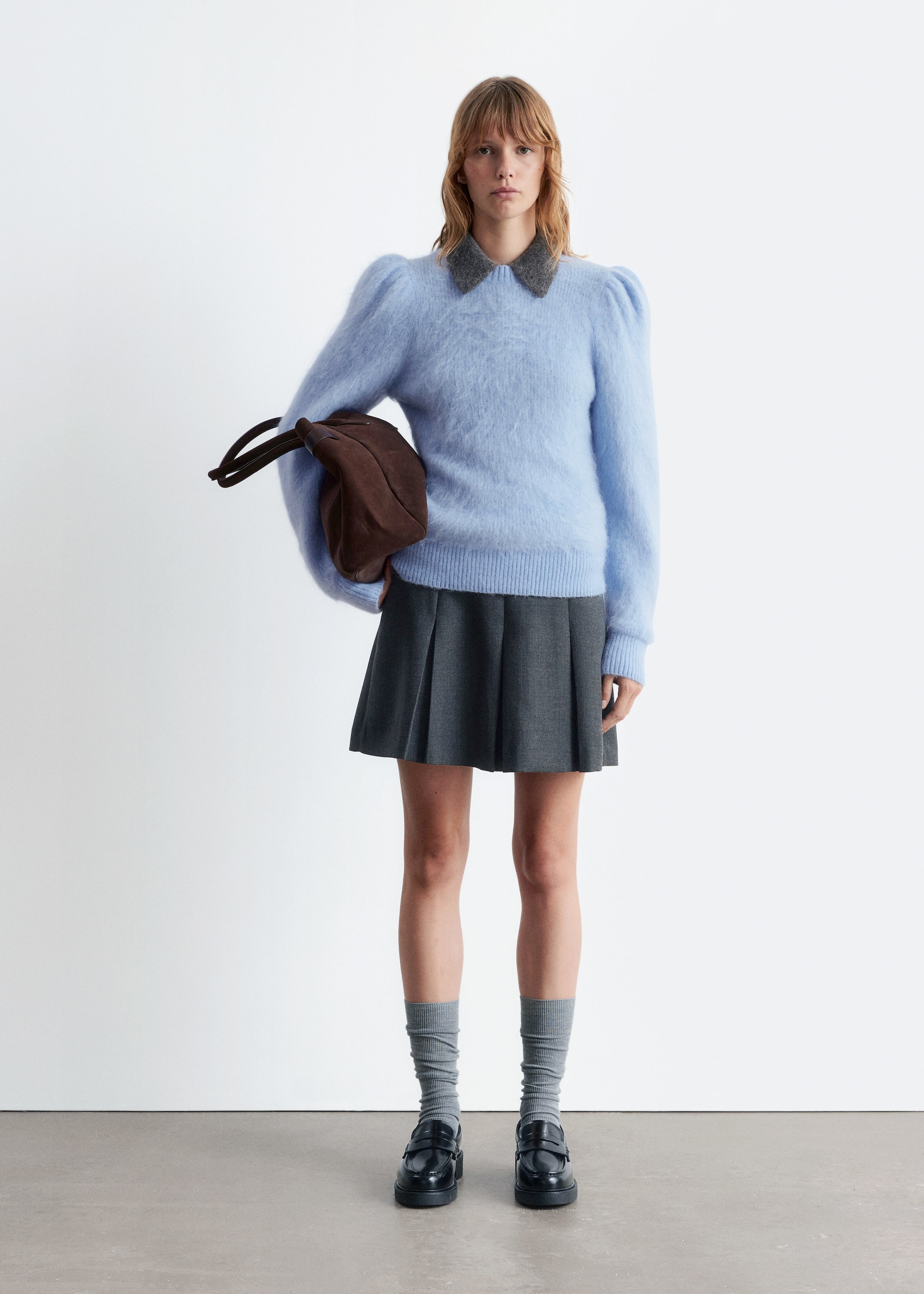 Visa större bild: En ljusblå luddig tröja med puffade axlar och en grå texturerad krage bärs tillsammans med en mörkgrå plisserad minikjol. Grå ribbade strumpor bärs med svarta chunky loafers, och en brun mjukstrukturerad väska hålls.