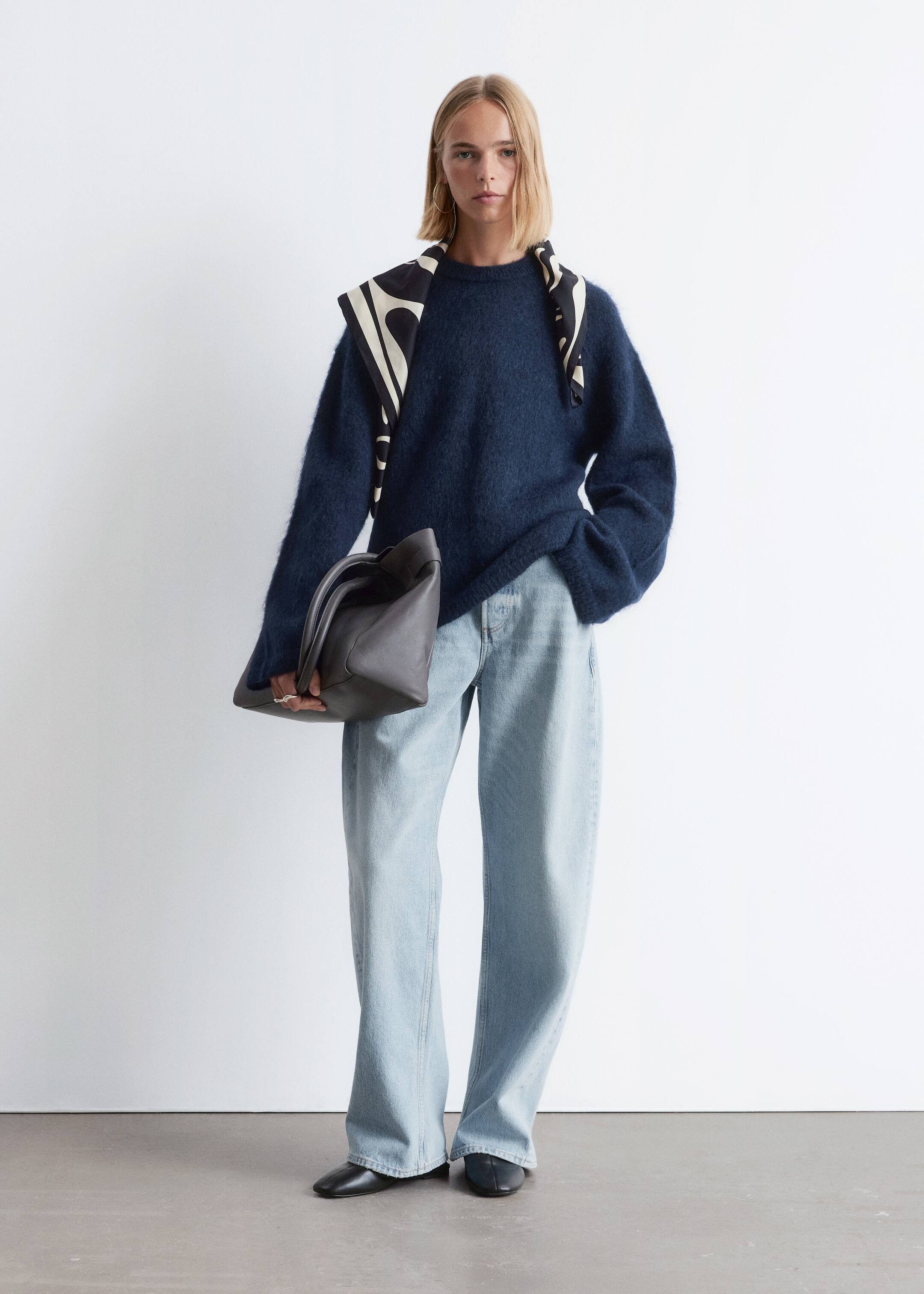 Agrandir l'image: Un mannequin se tient debout, face à l'avant, portant un pull à col rond bleu marine à coupe décontractée et à texture duveteuse, associé à un jean à jambes larges et délavé clair. Une écharpe à motifs noir et blanc cassé est drapée sur les épaules, et un sac à main structuré gris foncé est tenu, avec des chaussures plates à bout pointu noires visibles.