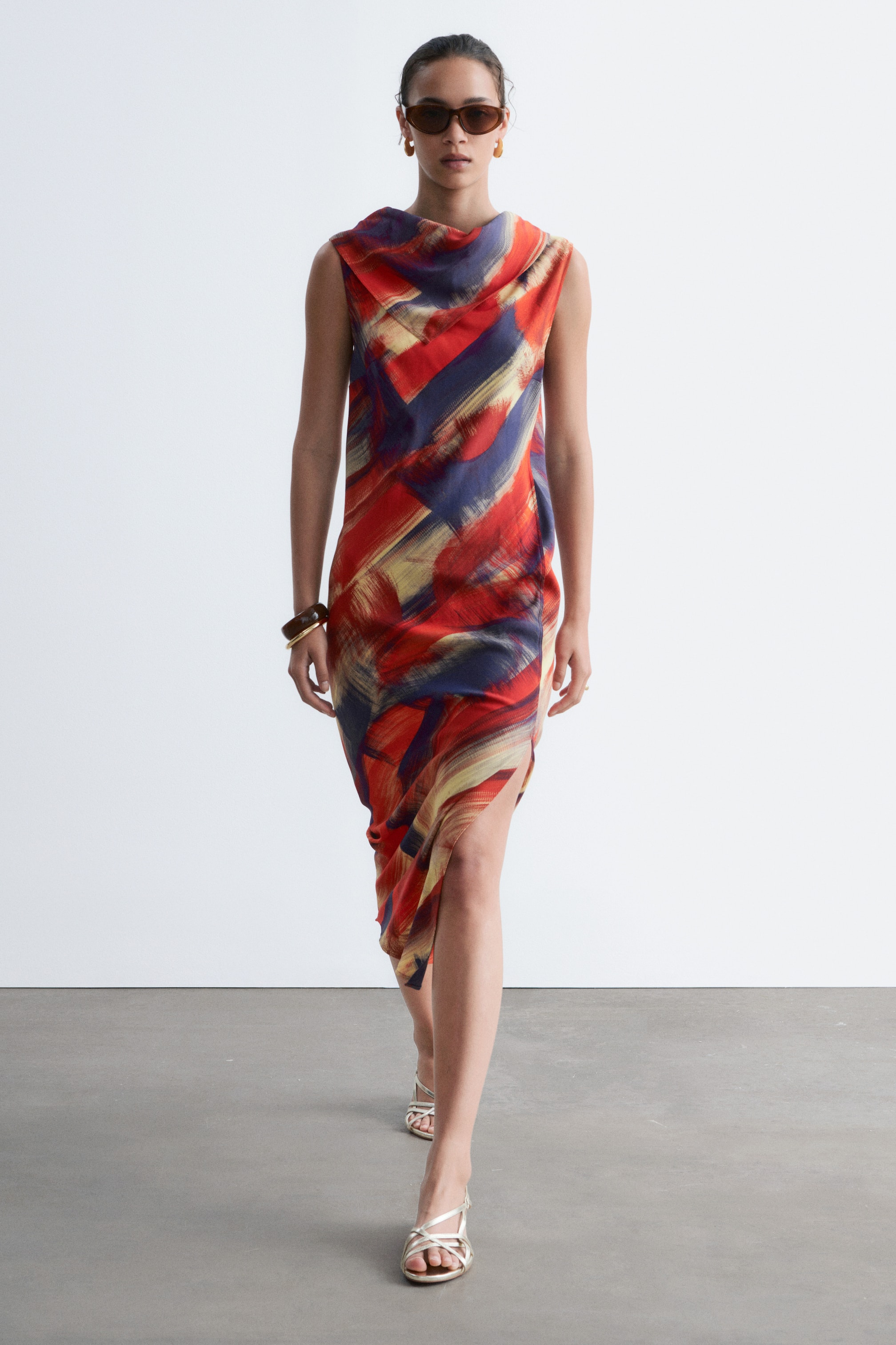 Grösseres Bild ansehen: Ein ärmelloses Kleid mit einem drapierten Wasserfallausschnitt und einem abstrakten Muster aus roten, blauen und gelben Pinselstrichen wird vom Model getragen. Die geschwungenen Seitennähte erzeugen einen subtilen Frontschlitz, und das Model läuft, während es goldene Sandalen trägt.