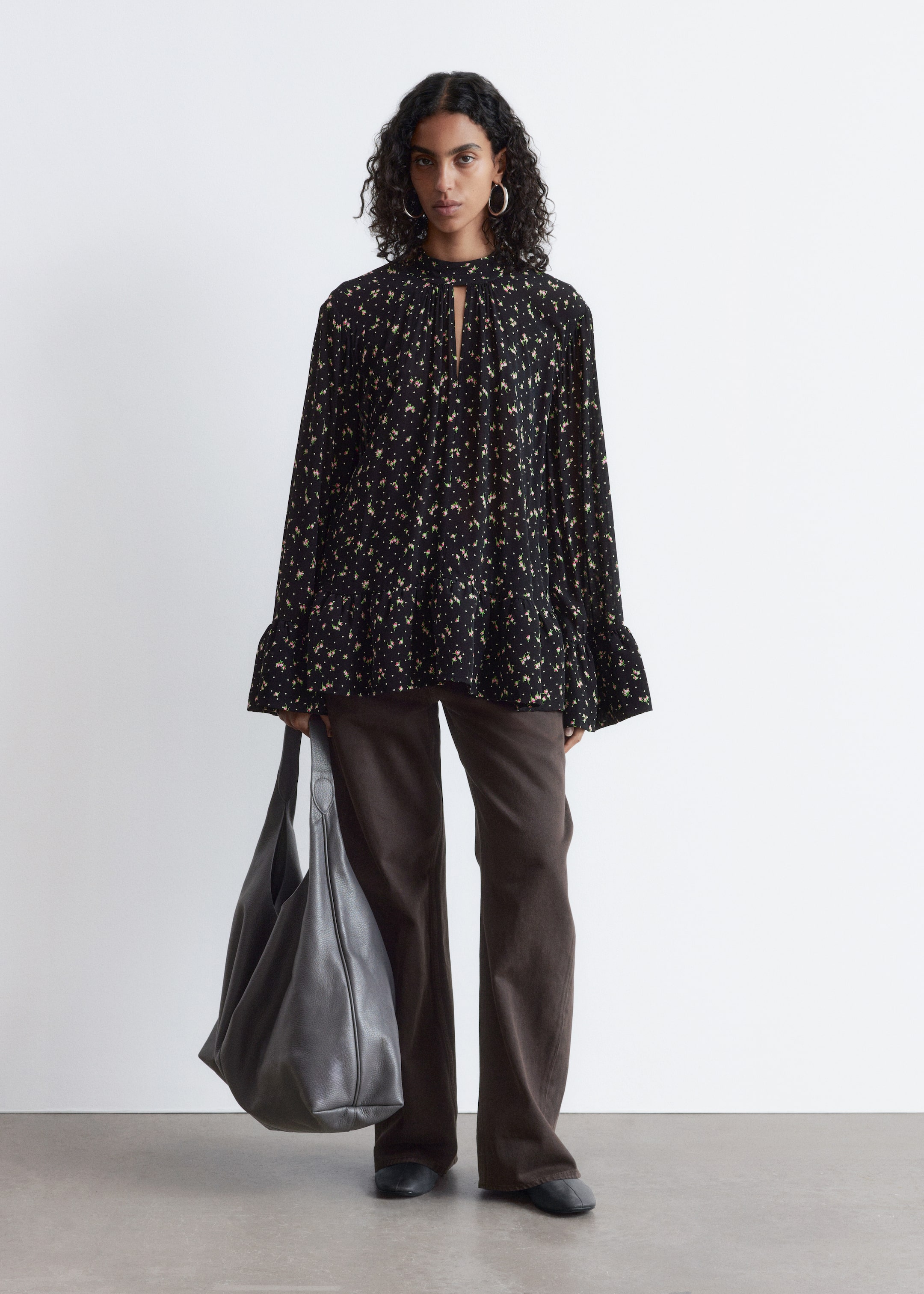 Agrandir l'image: Une blouse florale noire présente une coupe ample et décontractée, orientée vers l'avant. Elle arbore un col haut avec une découpe en trou de serrure et des fronces, des manches longues et larges avec des poignets froncés, ainsi qu'un ourlet évasé, orné d'un motif floral jaune, rose et blanc.