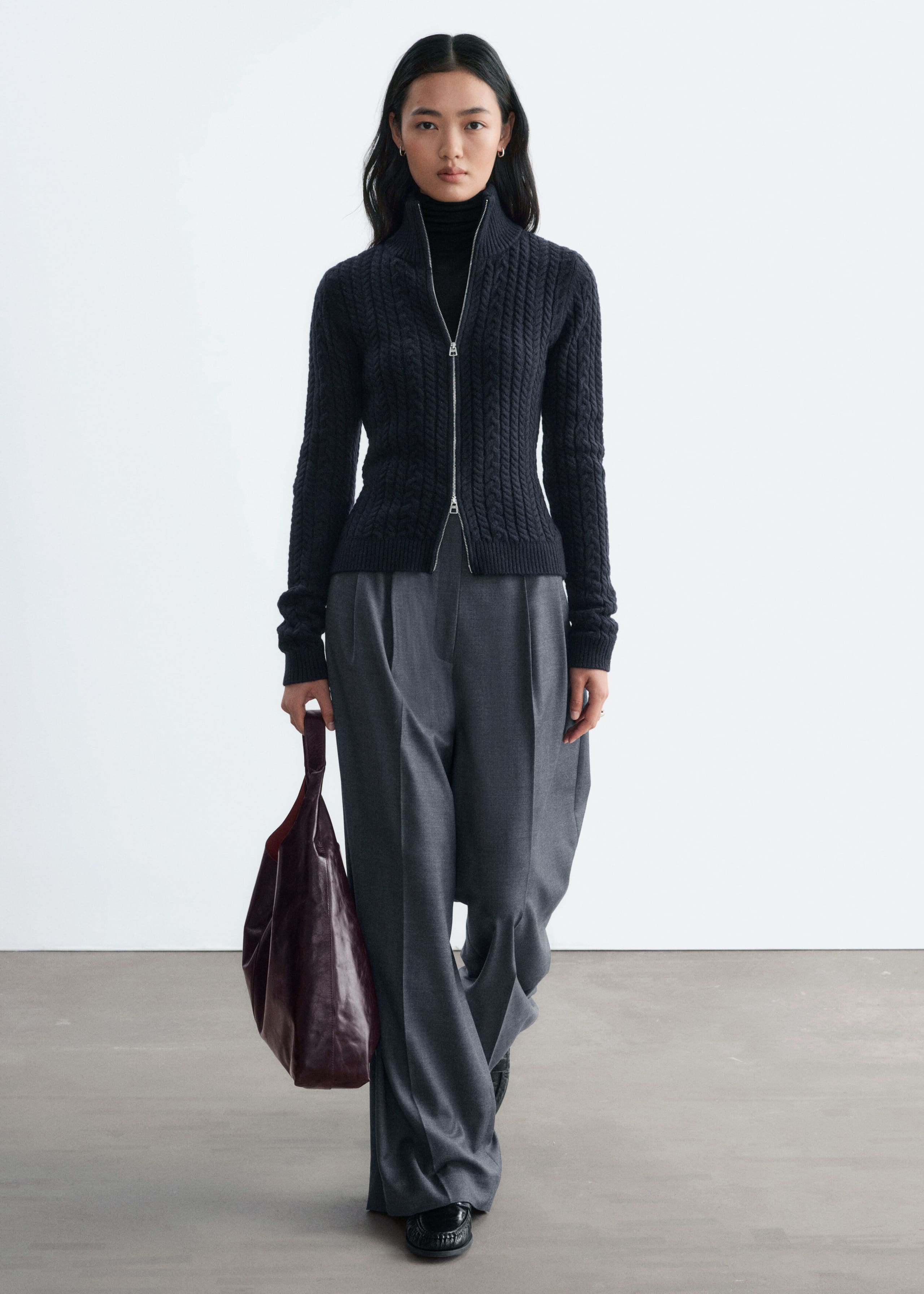 A slim-fit, dark navy cable-knit cardigan is being worn by the model, featuring a double-zip front closure and a high ribbed collar. The cardigan is layered over a black roll-neck and paired with wide-leg grey trousers, while a dark burgundy leather bag is being held in one hand.