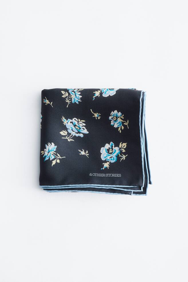 A dark grey pocket square showcases a scattered floral pattern in light blue and white with subtle yellow details, and a light blue trim outlines its folded edges. The "& OTHER STORIES" logo appears in white on the fabric. - 1
