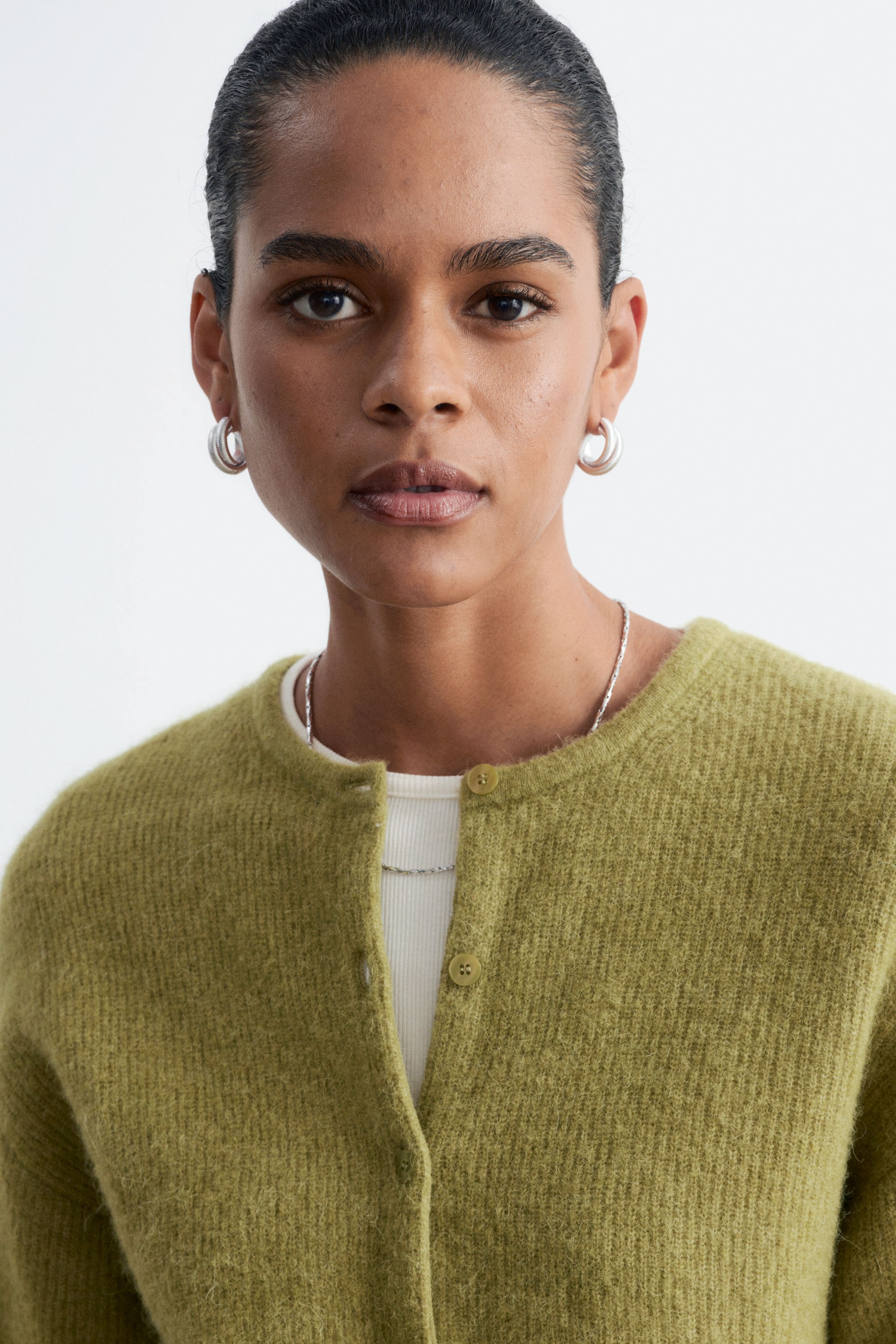 Größeres Bild ansehen: Ein grüner Strick-Cardigan wird vom Model über einem weißen gerippten Oberteil getragen. Der Cardigan zeichnet sich durch eine flauschige Struktur, einen Rundhalsausschnitt und Knopfleisten vorne aus, wobei die oberen Knöpfe geschlossen sind.
