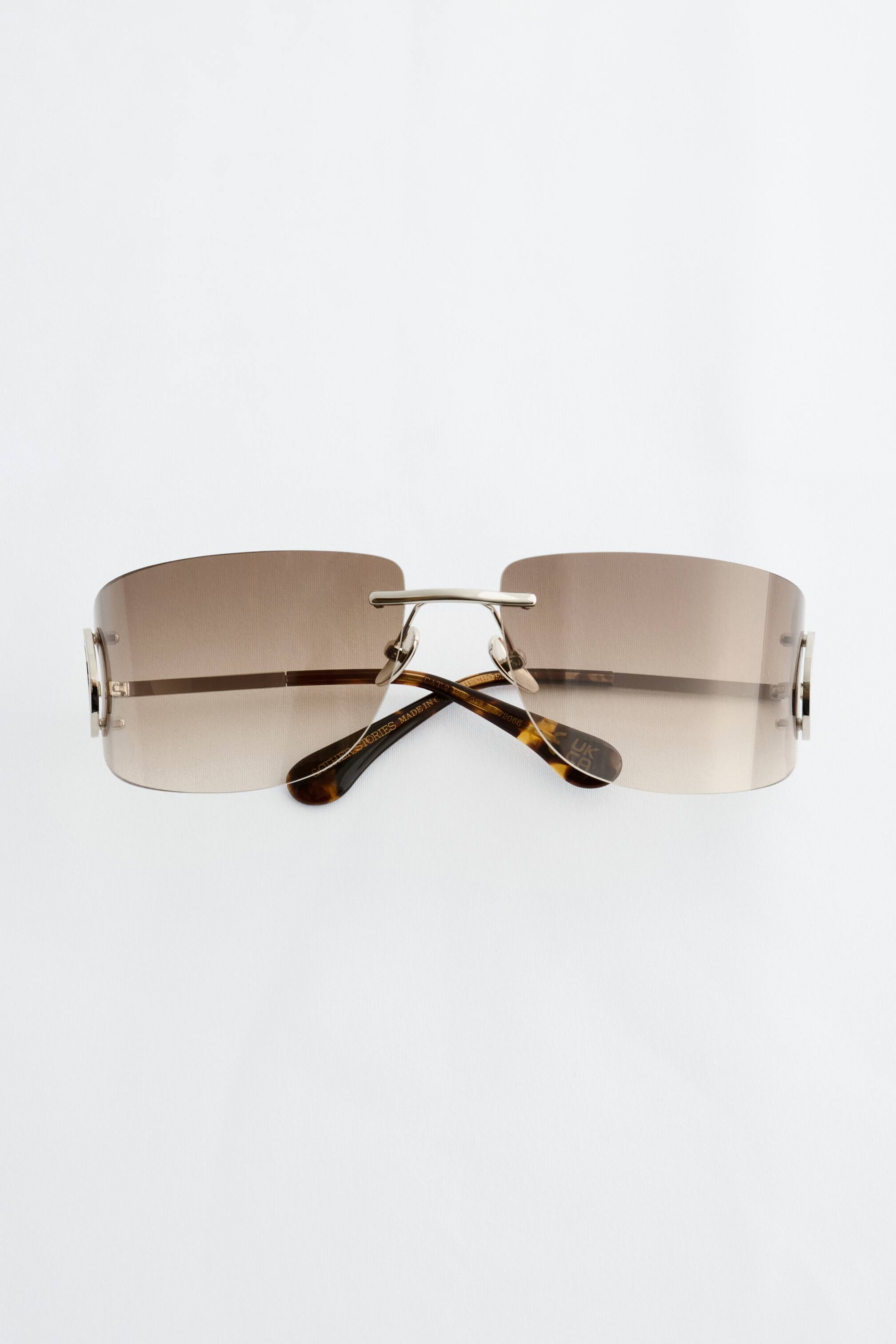 Größeres Bild ansehen: Die Sonnenbrille präsentiert randlose, umschließende, sanft rechteckige Gläser in einem warmen braunen Farbverlauf, die nach vorne ausgerichtet sind. Runde, silberfarbene Bügeldetails verleihen dem Design einen von Vintage inspirierten Charakter, mit Schildpattspitzen, die an den gefalteten Bügelarmen sichtbar sind.