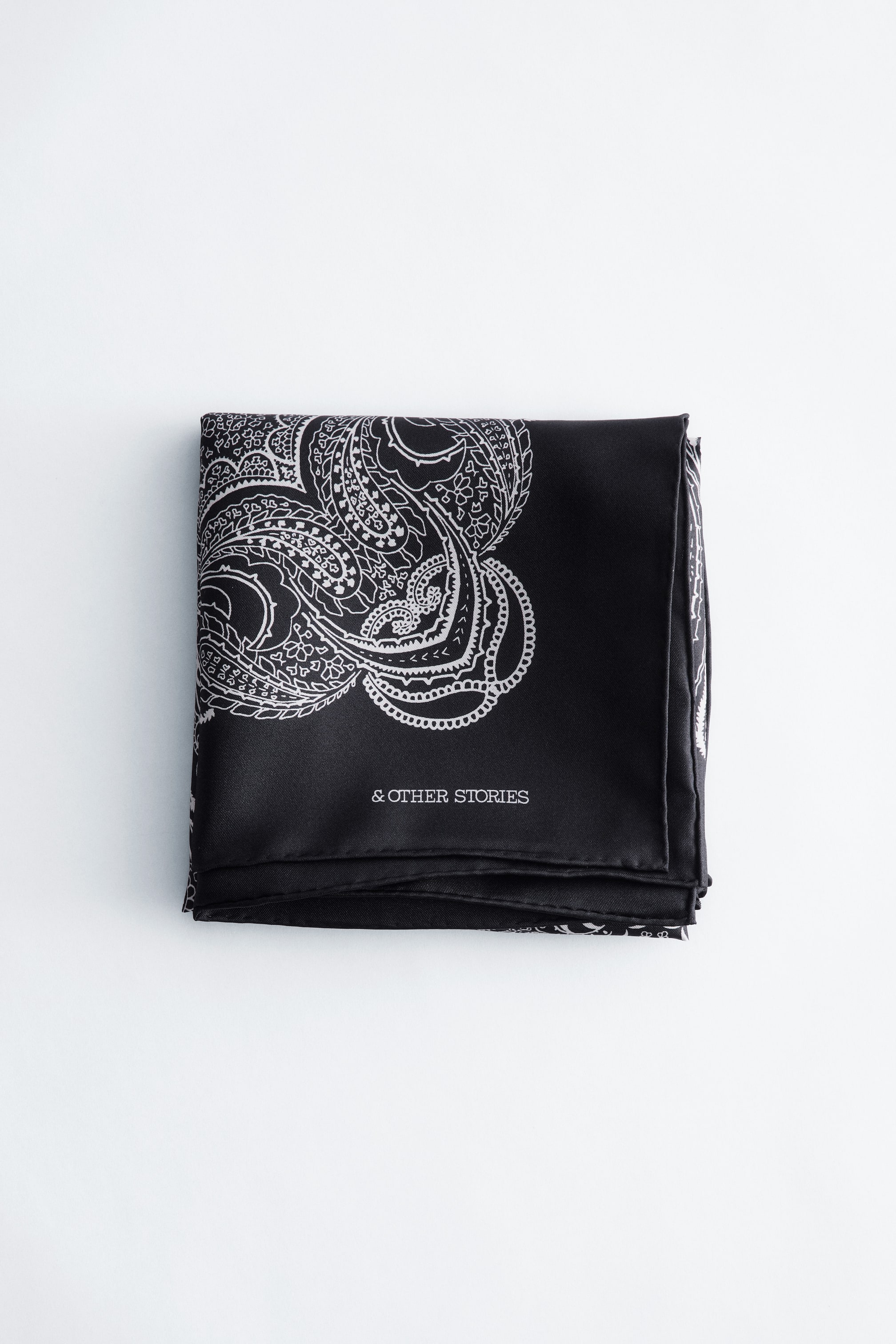 Zobrazit větší obrázek: Černý hedvábný šátek je úhledně složený do čtverce a na jeho povrchu se rozprostírá velký bílý paisley vzor. Název značky '& OTHER STORIES' je na šátku vytištěn bílým písmem.
